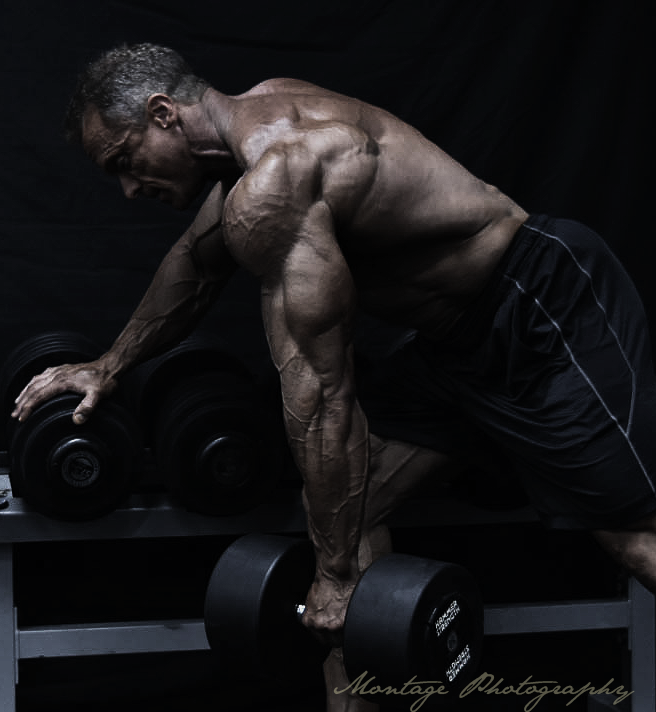 Muscular athlete performing dumbbell row exercise in dramatic low-key lighting.