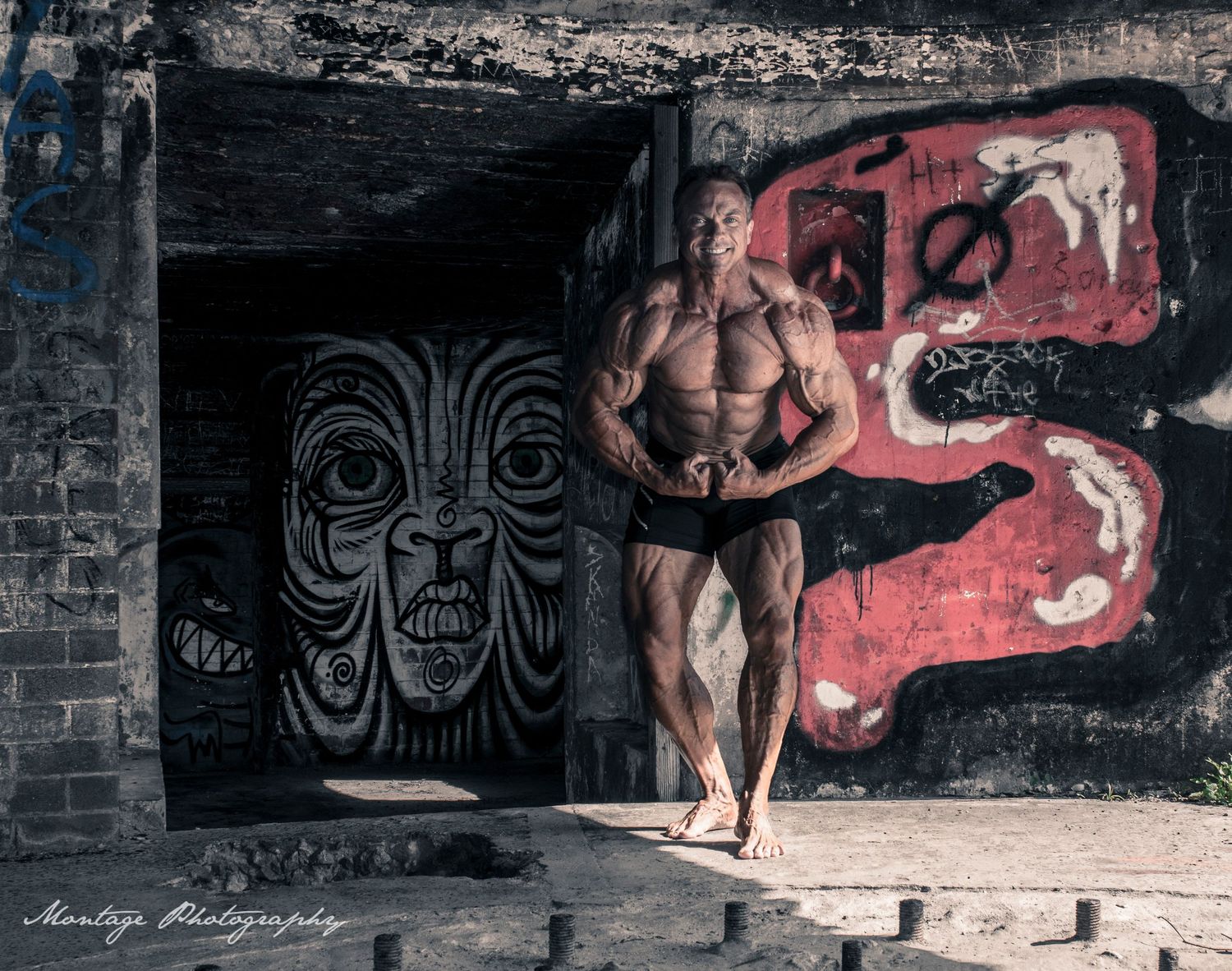 Muscular bodybuilder posing against urban graffiti wall with tribal mask artwork in dark atmospheric setting.