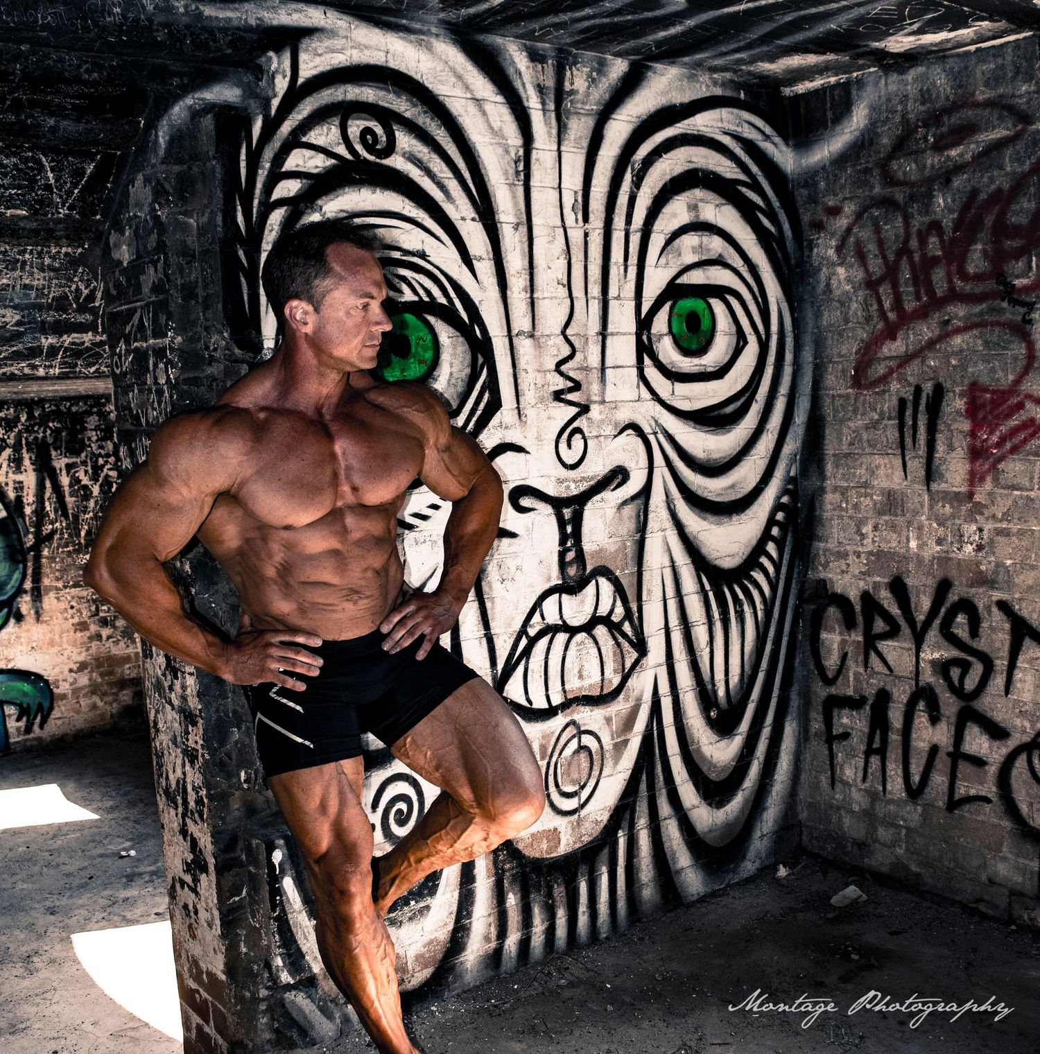 Muscular athlete poses against urban wall art featuring large black and white face with striking green eyes painted as graffiti.