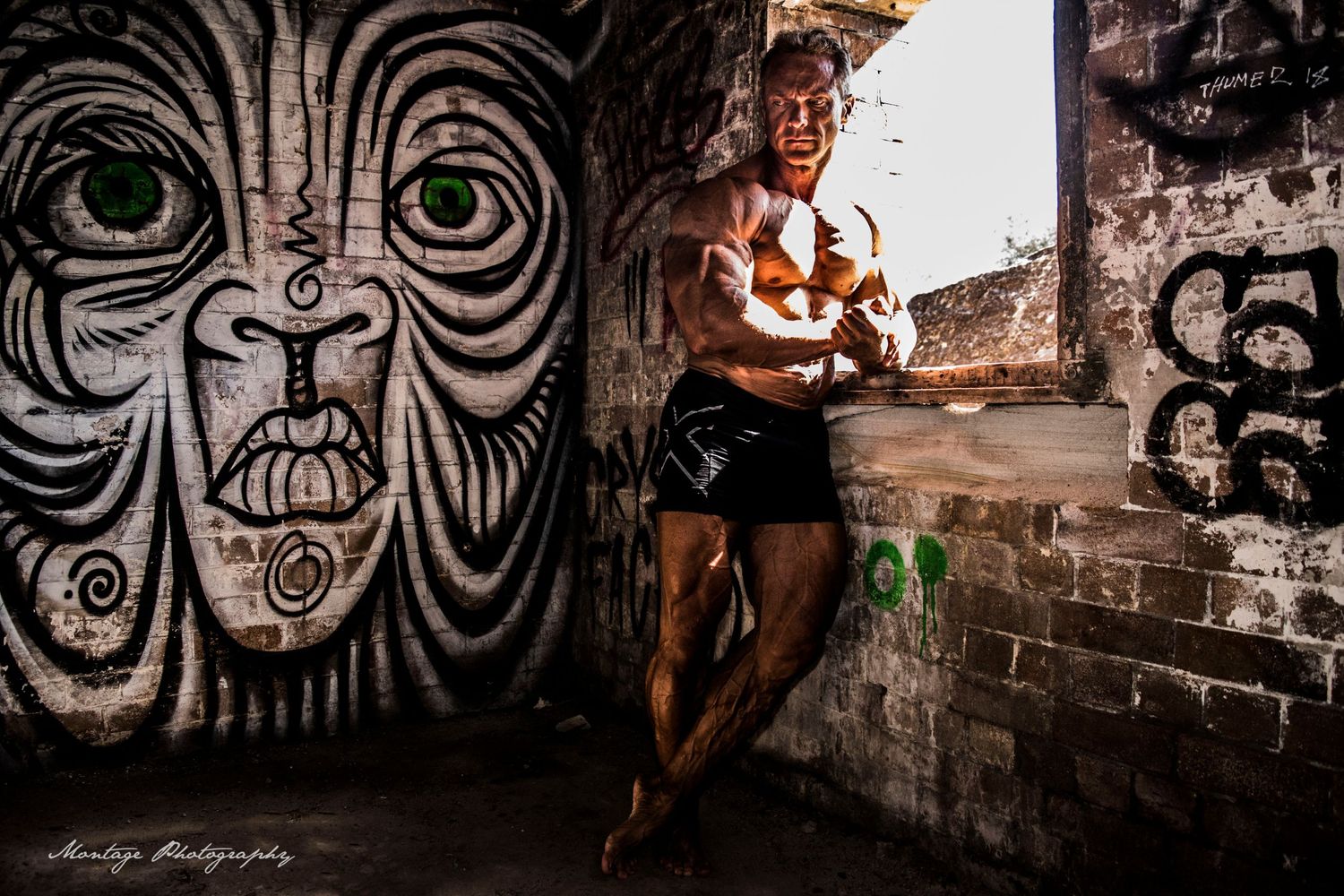Athletic physique displayed against brick wall with dramatic tribal face graffiti artwork backdrop.