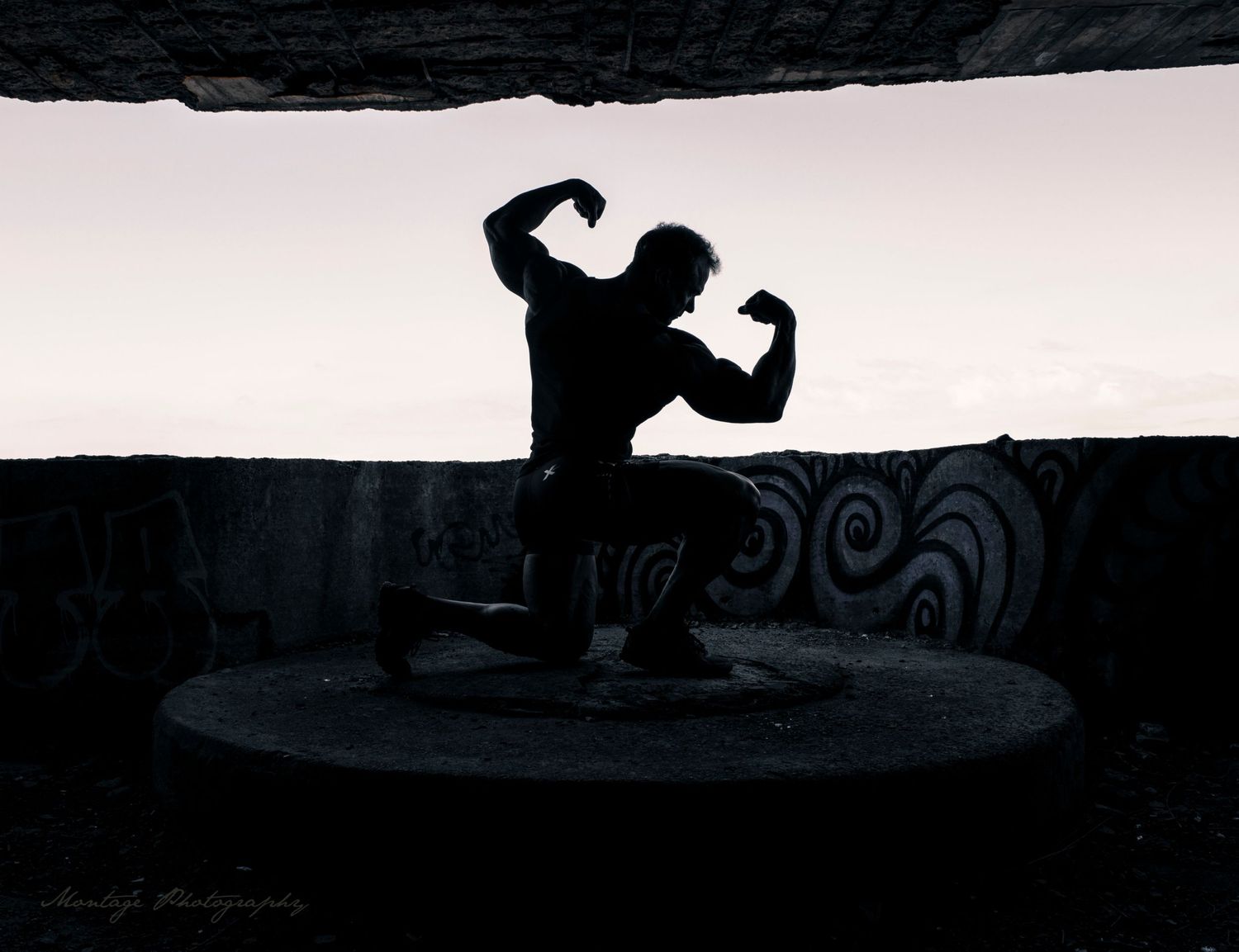 Dramatic silhouette of bodybuilder posing on circular platform against light background.