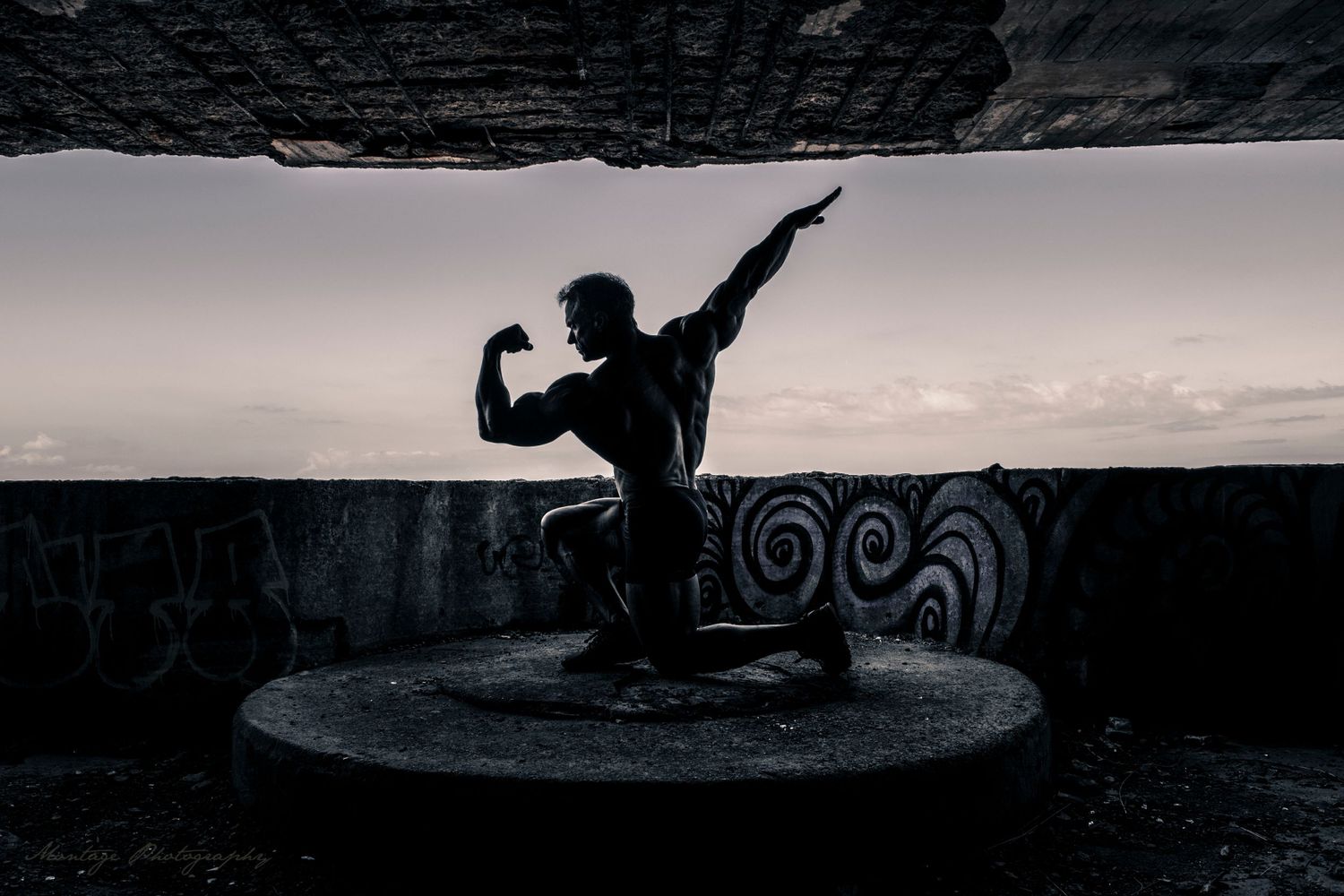 Silhouette of bodybuilder performing graceful pose against dramatic sky beneath stone archway in black and white.