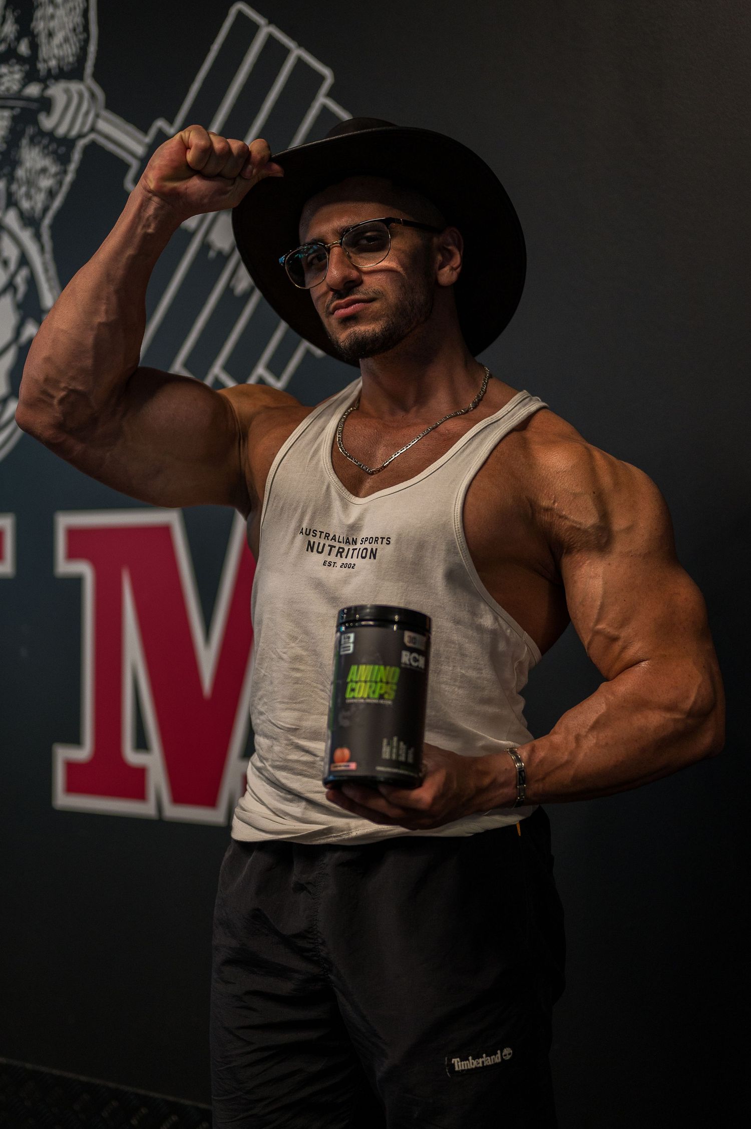 Bodybuilder in hat and tank top posing with supplement container against gym wall.