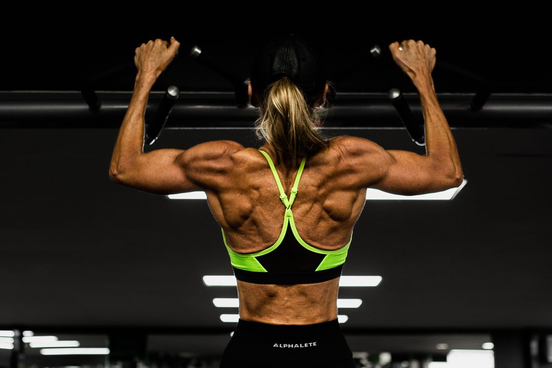 Muscular back view of person exercising at gym wearing neon green sports bra showing defined muscles.
