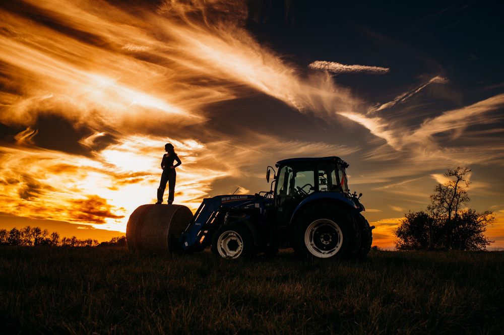 Silhouette of figure standing on tractor against dramatic orange sunset sky with wispy clouds.