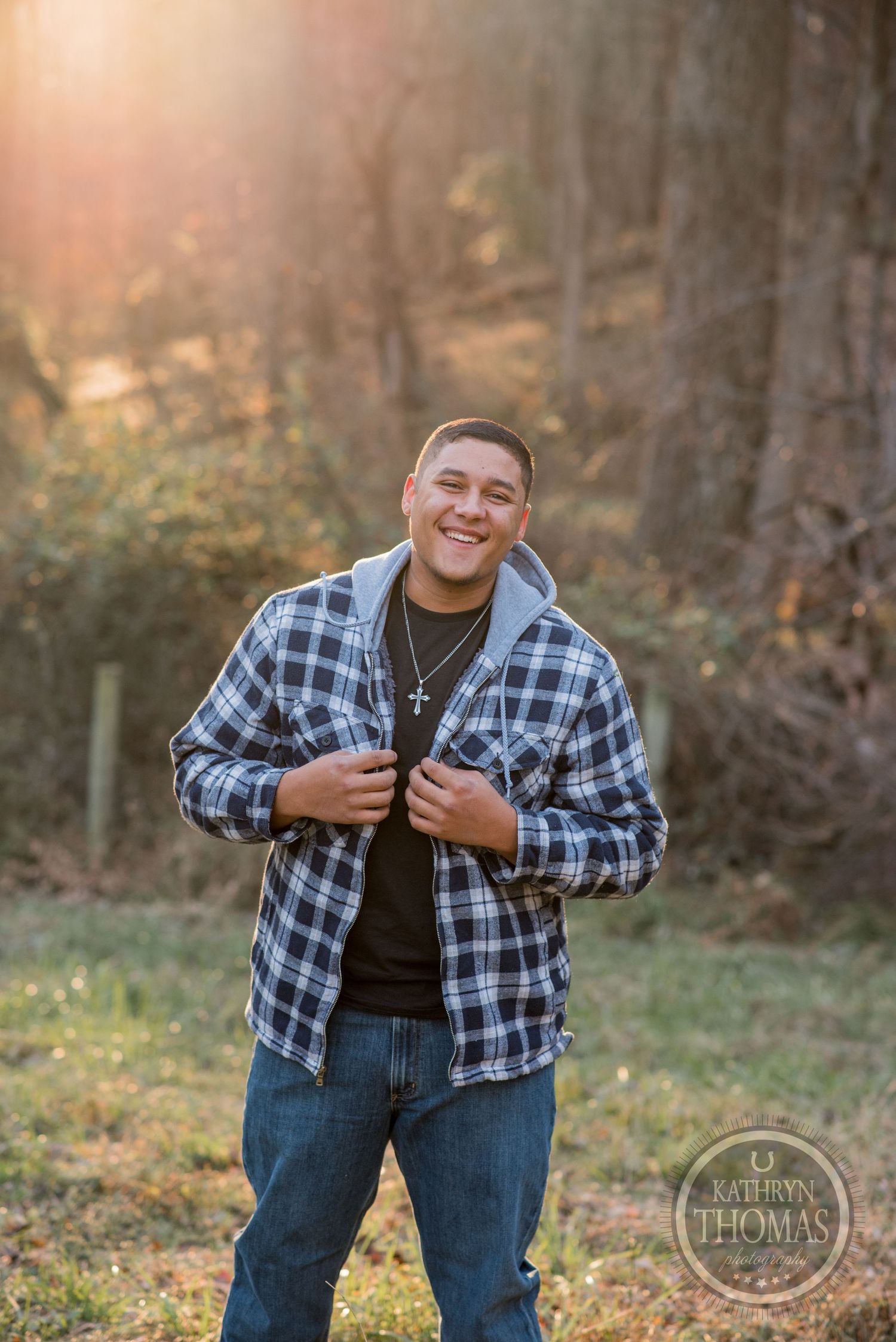 Someone in a black and white plaid shirt and cross necklace smiles in a sunlit forest setting.