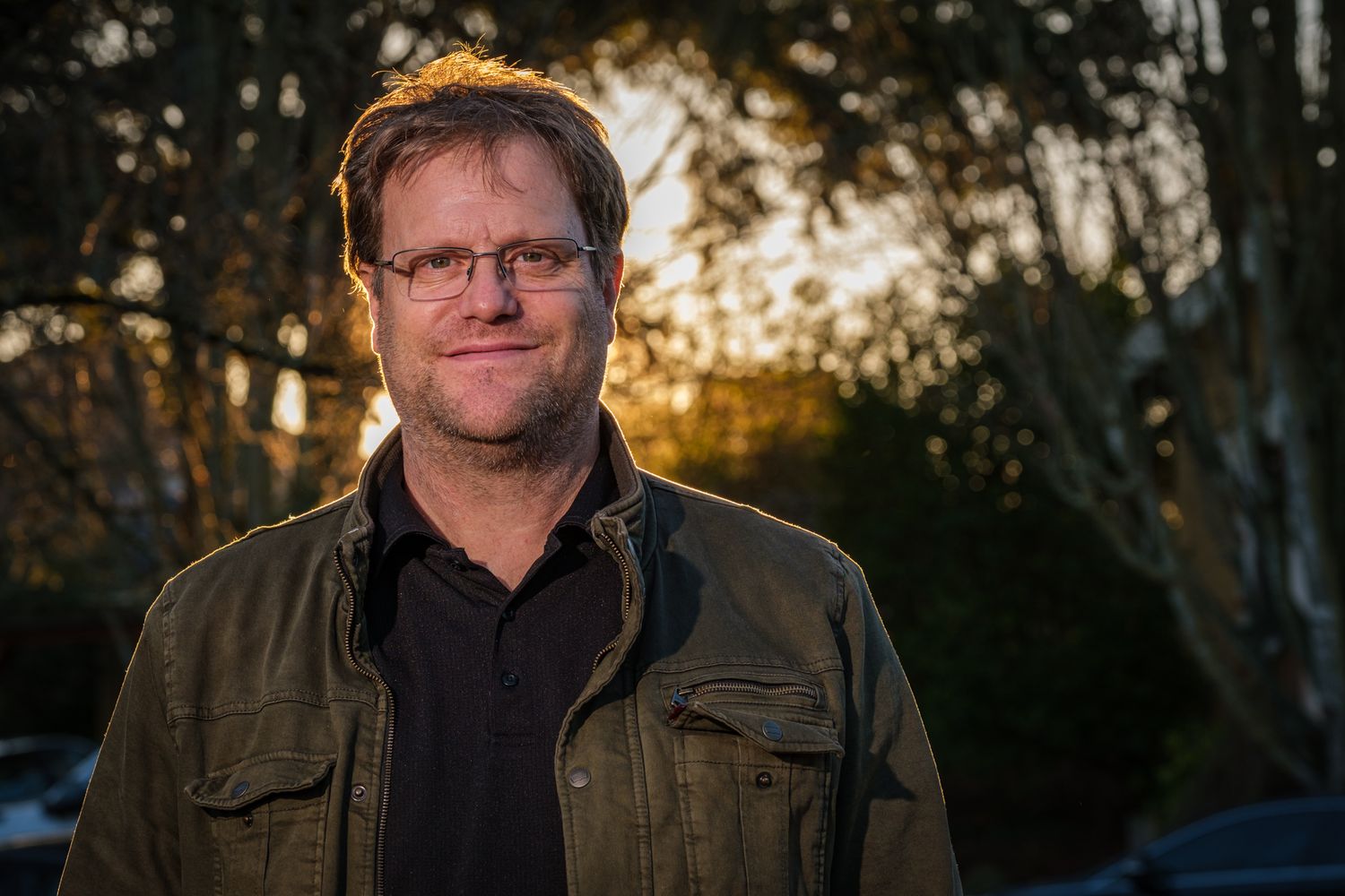 A person wearing a brown jacket stands outdoors at sunset with trees in the background.