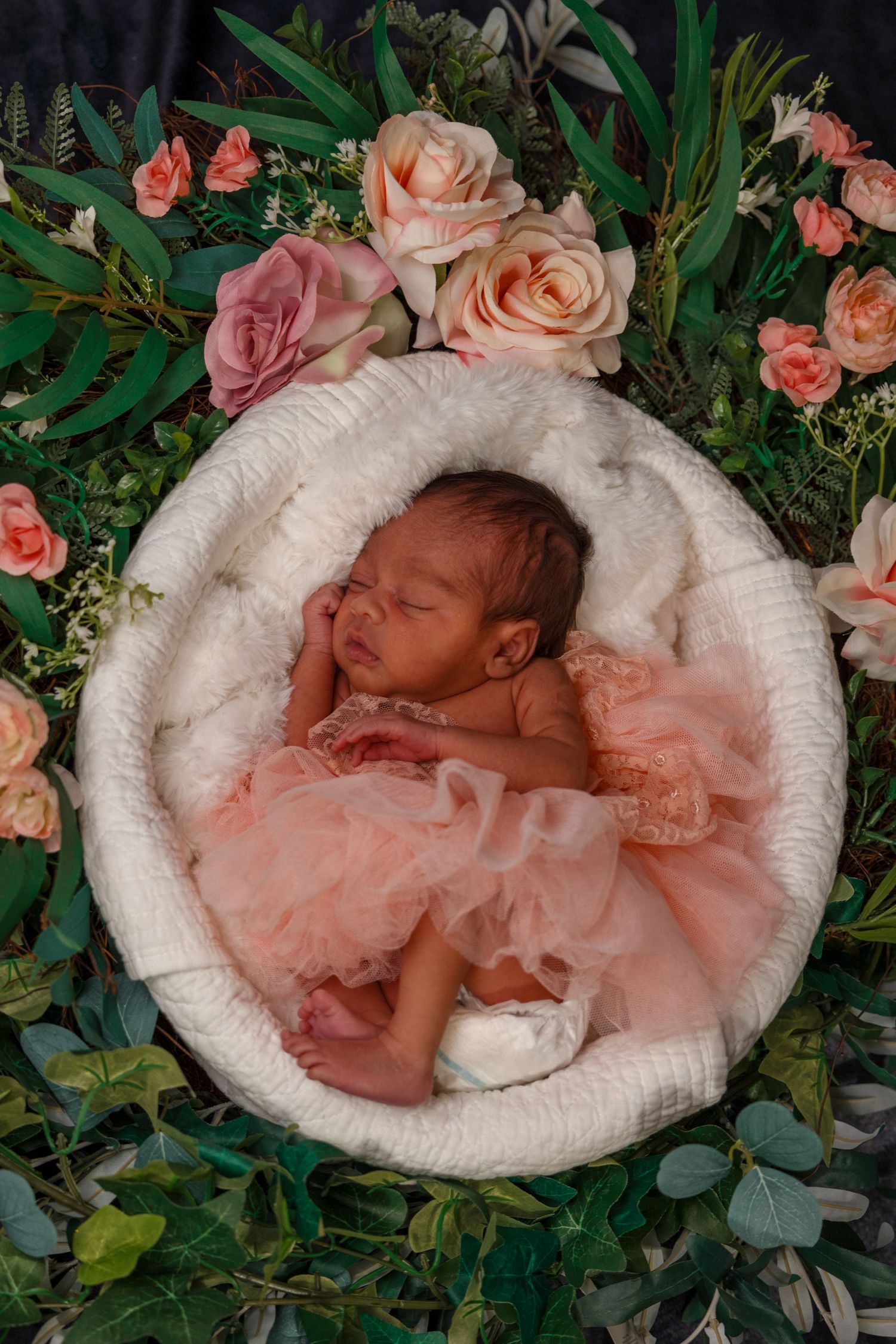 Sleeping newborn in pink tulle dress nestled in white basket with floral decorations.
