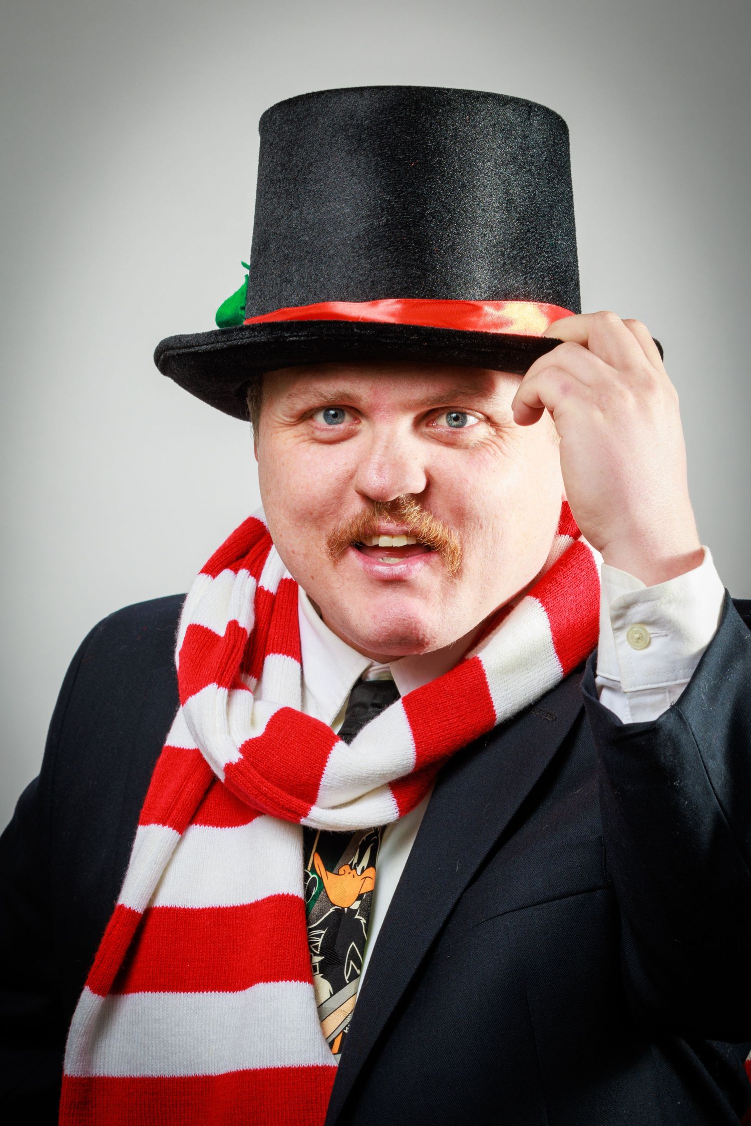 Person wearing a black suit and red and white striped scarf tips their top hat in a playful pose.