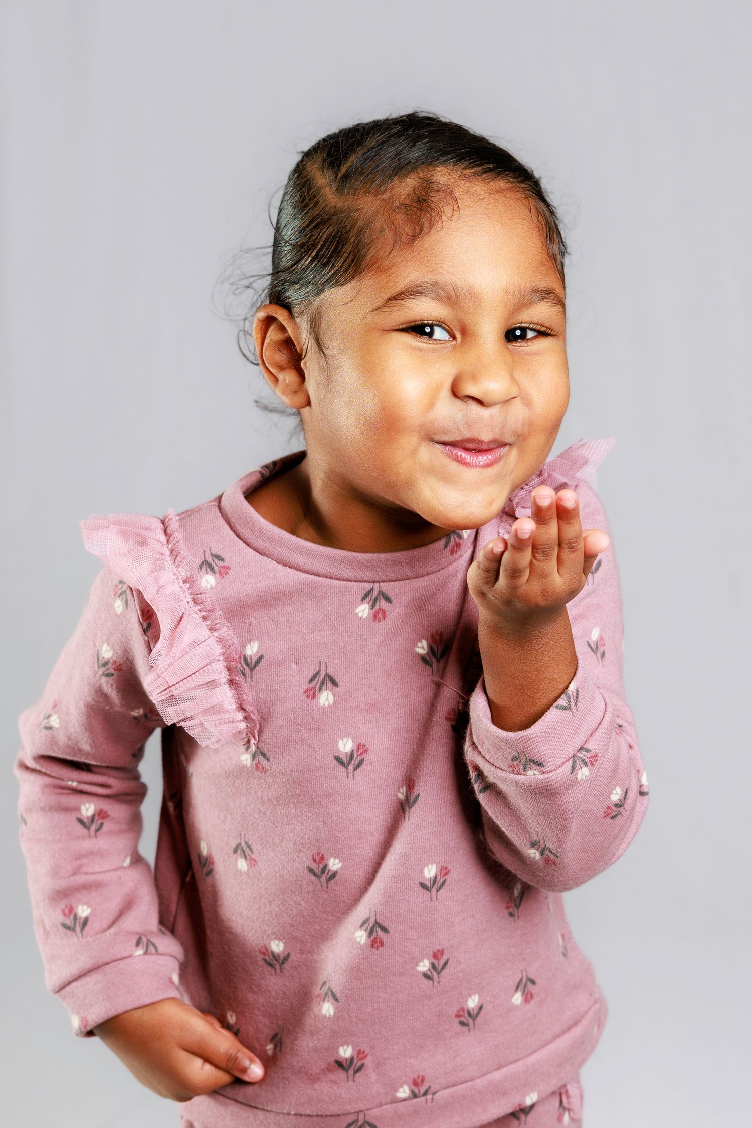 Child in pink ruffled sweater makes playful expressions in studio portraits against gray background.