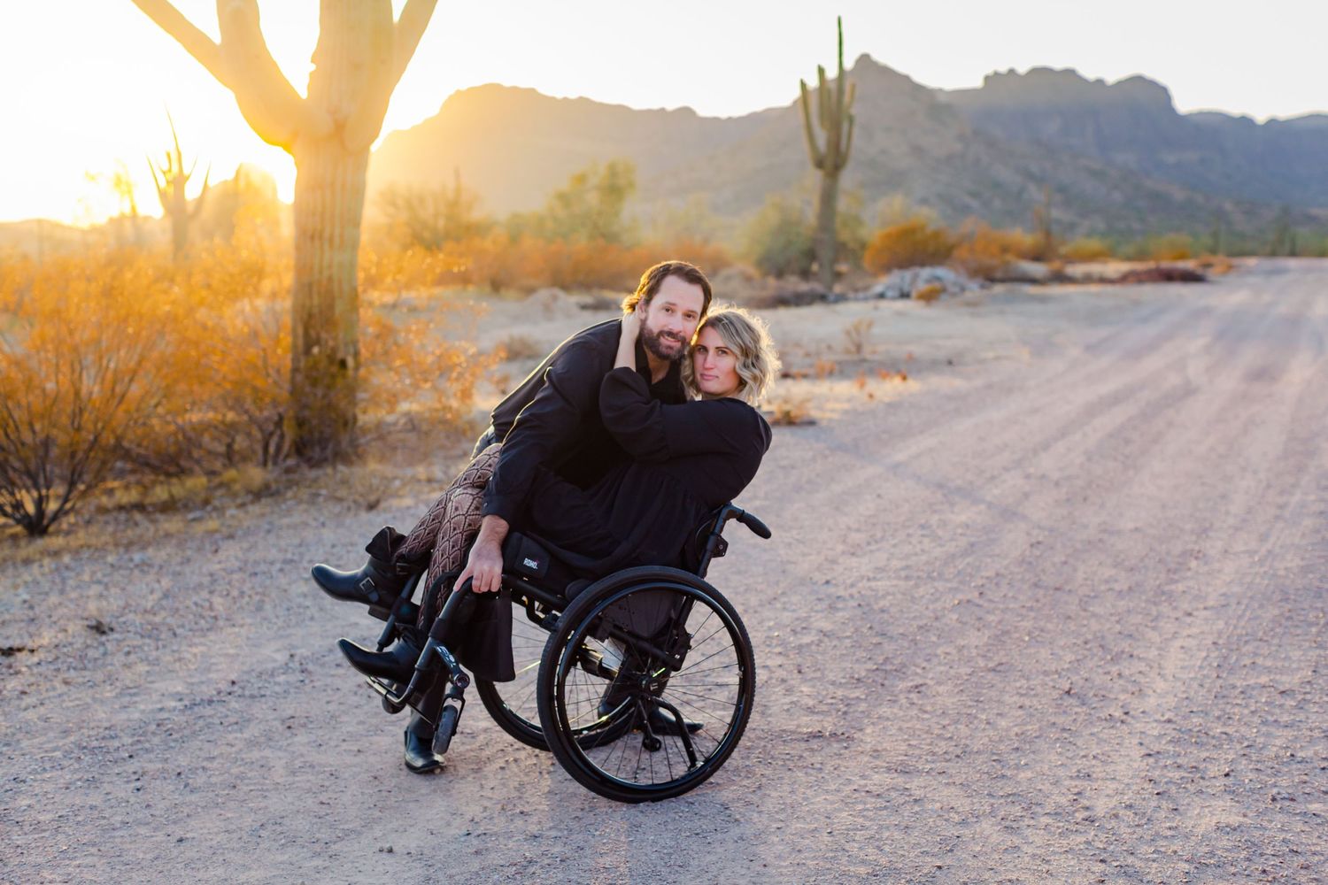 Couple shares tender moment on dirt road with desert mountains in background during sunset.
