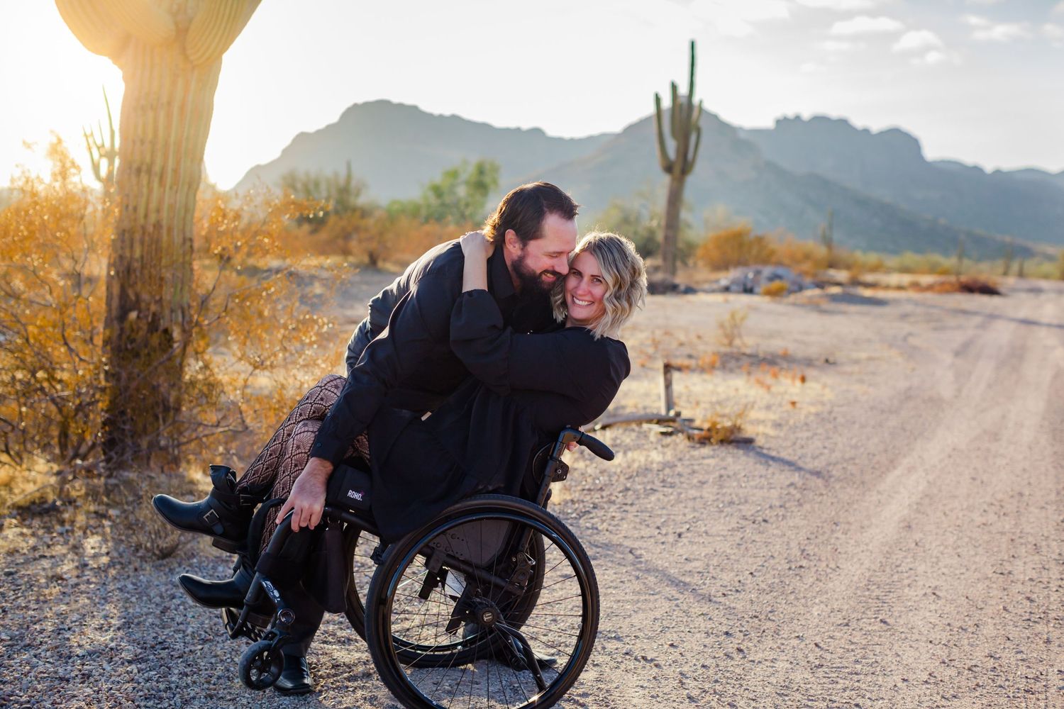 Wheelchair user and companion share intimate moment against desert mountain backdrop at sunset.
