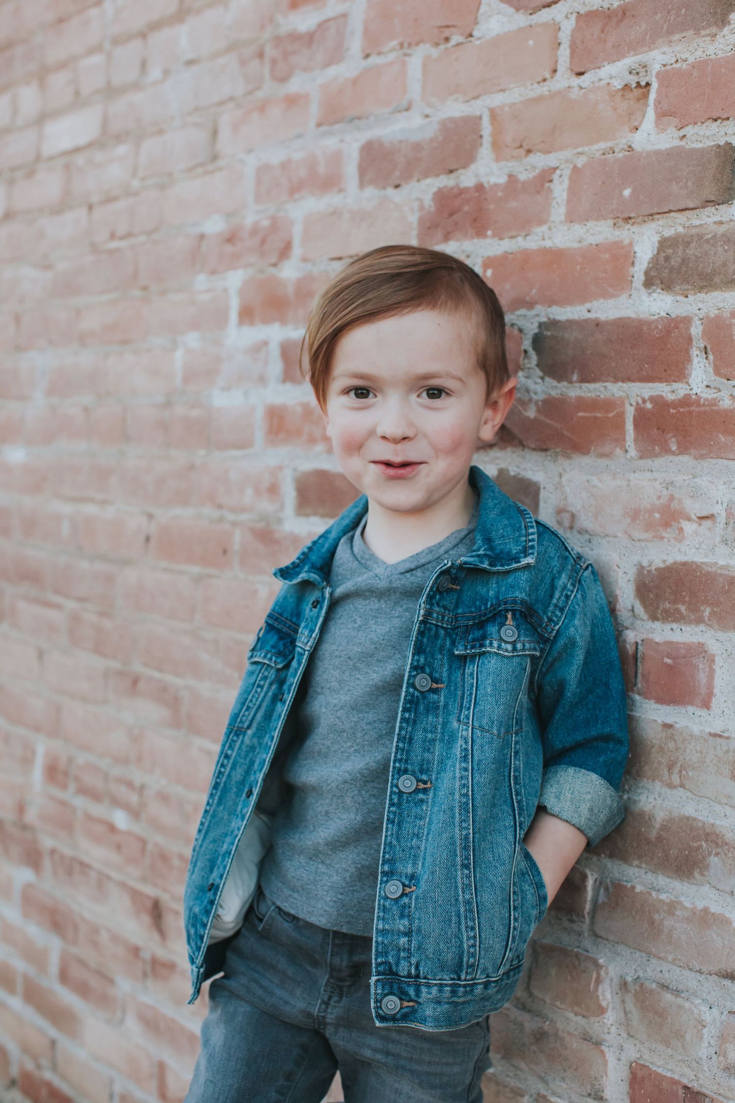 Child in denim jacket poses against weathered brick wall in natural lighting.