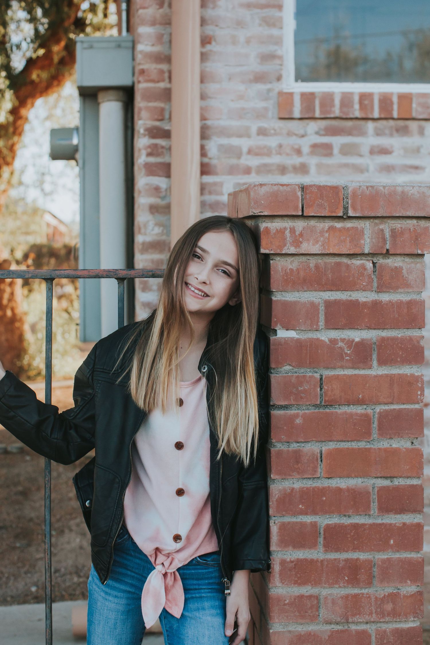 Woman in denim and black jacket poses against rustic brick wall outdoors.