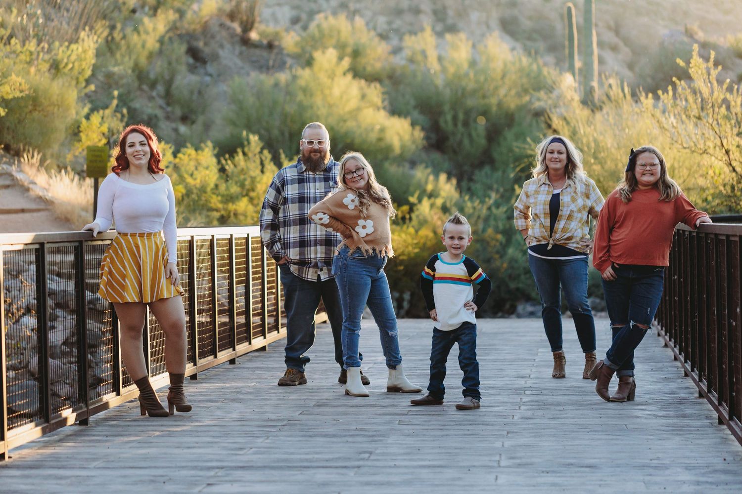 People walking and posing on a wooden boardwalk surrounded by trees at sunset.