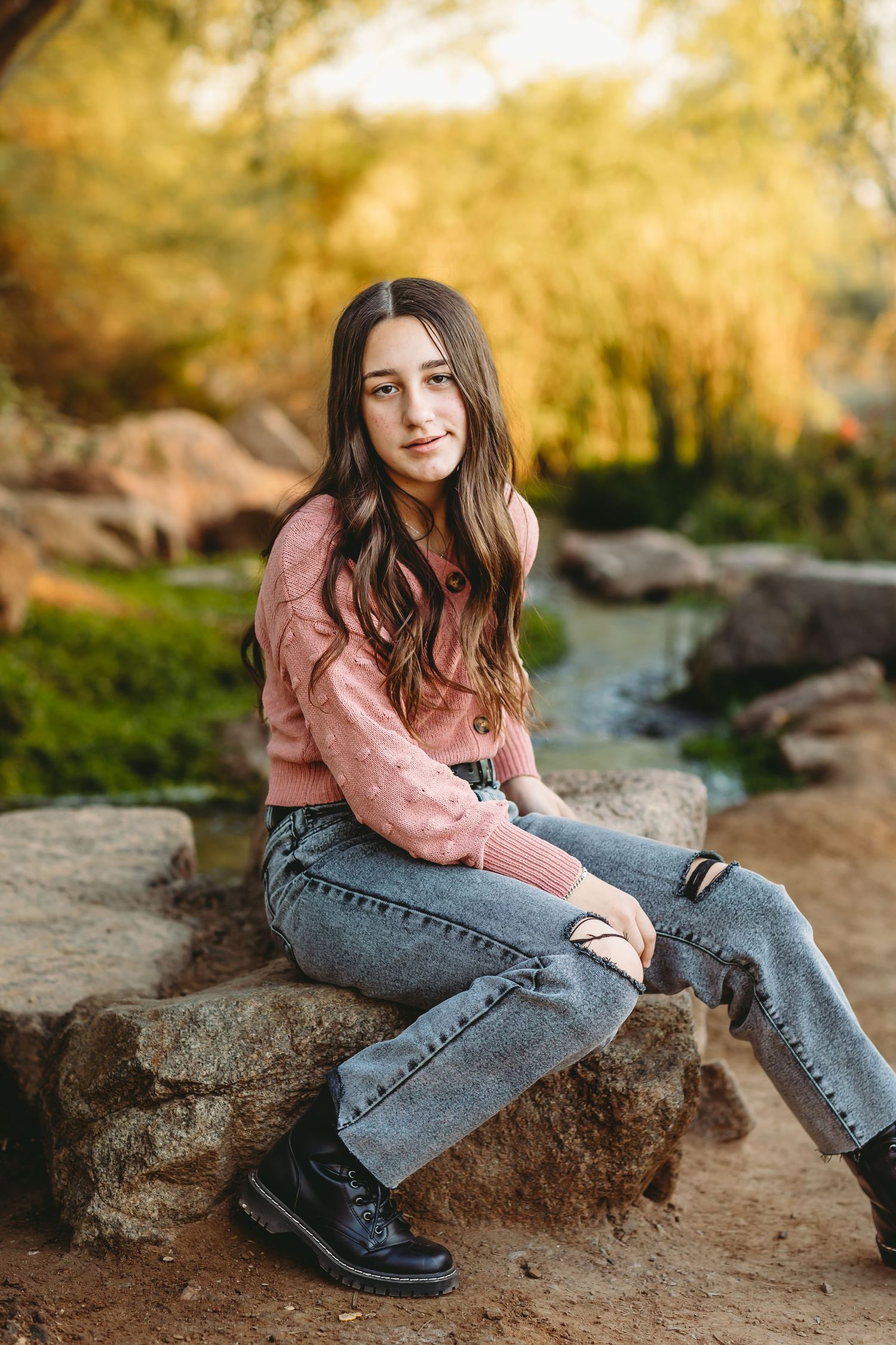 Person in pink sweater sits on rocks near stream with autumn foliage in background.