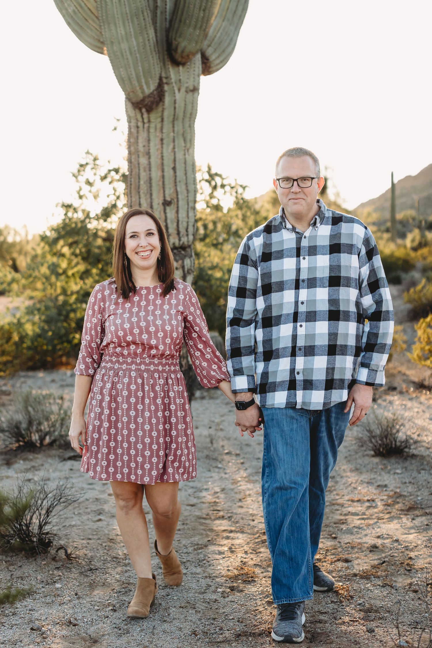 A couple stands together near a large saguaro cactus in the desert landscape during golden hour.