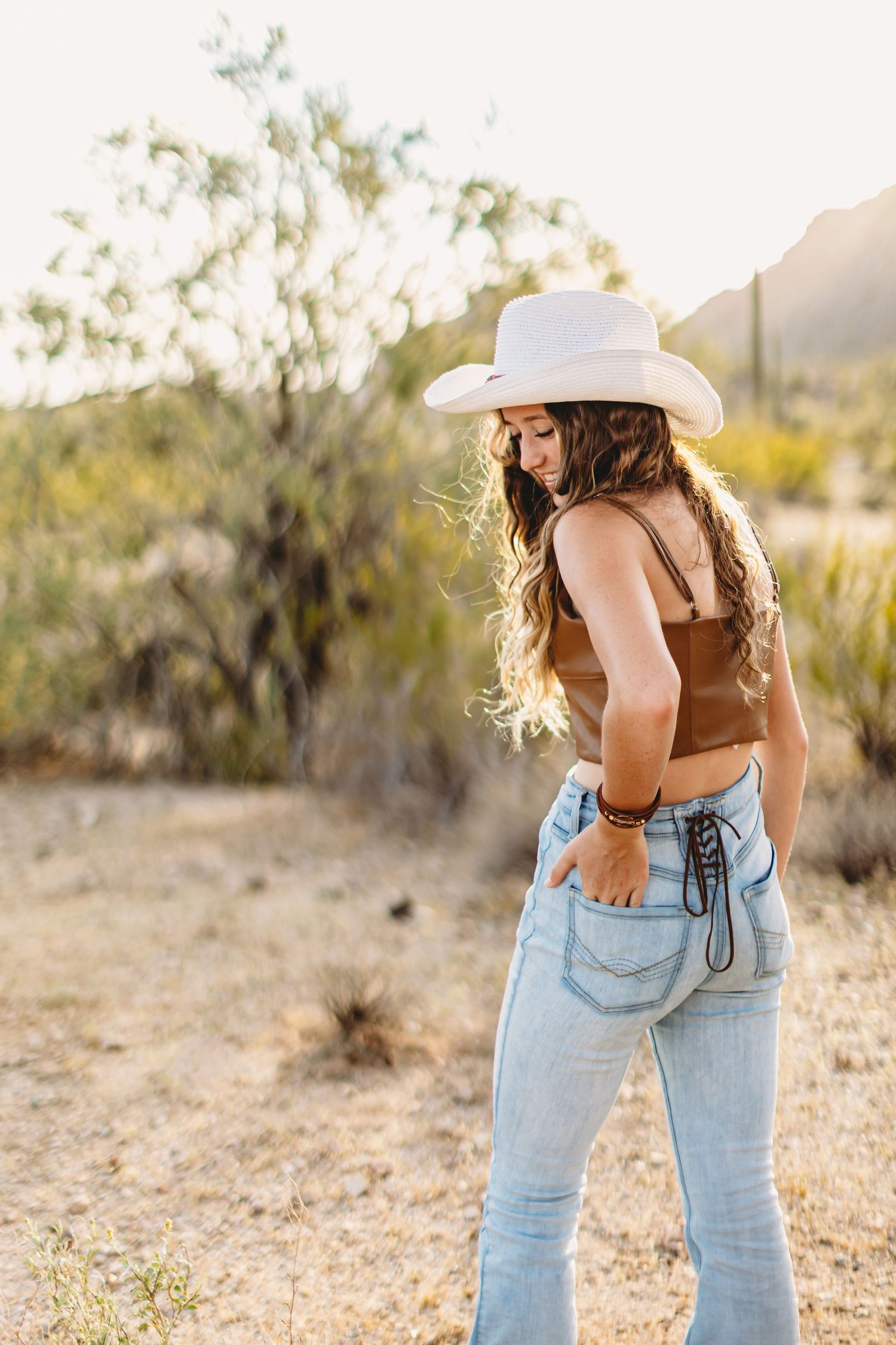 Woman in white cowboy hat and jeans poses against desert landscape at sunset.