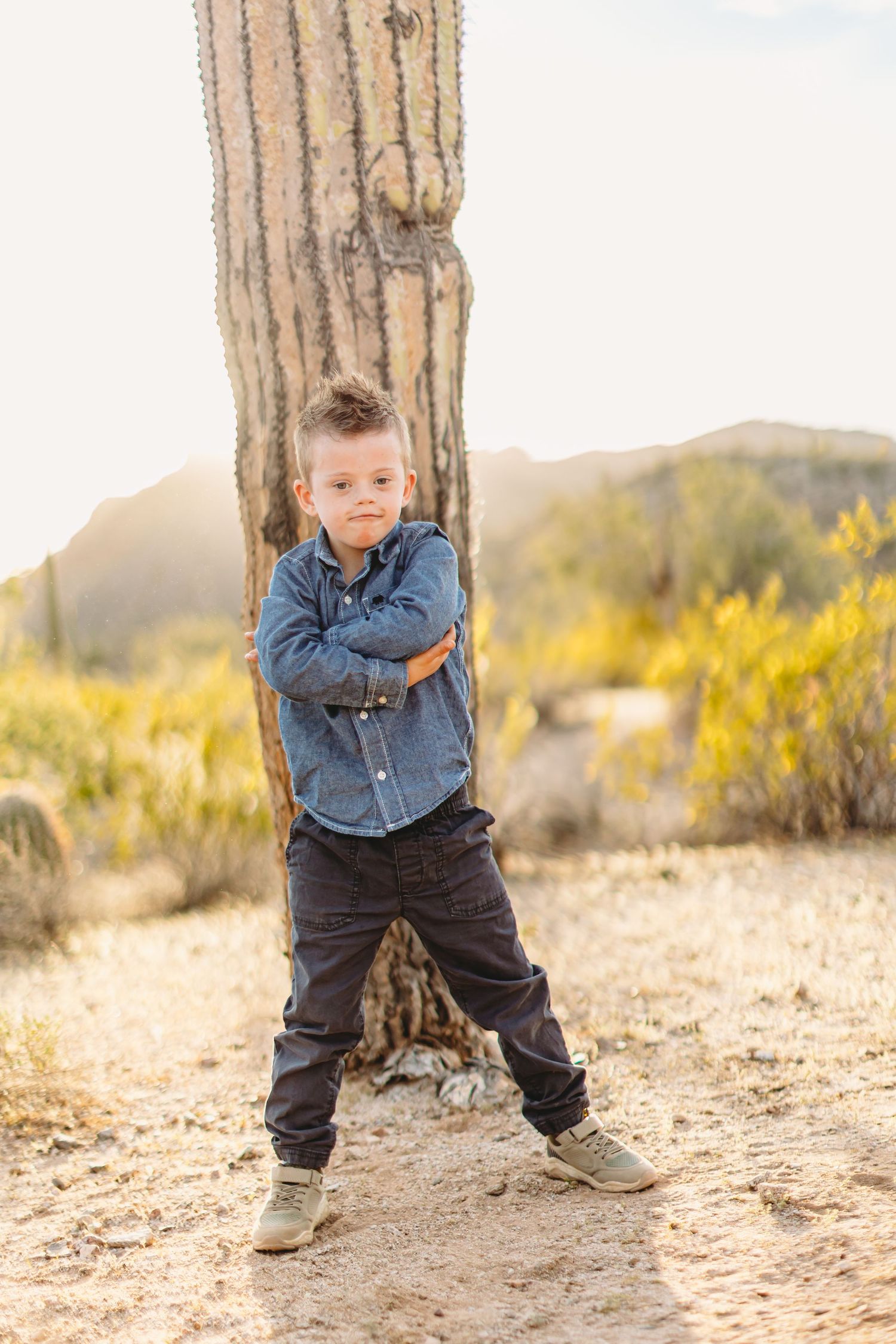 Young child in denim jacket poses playfully against saguaro cactus in desert setting.