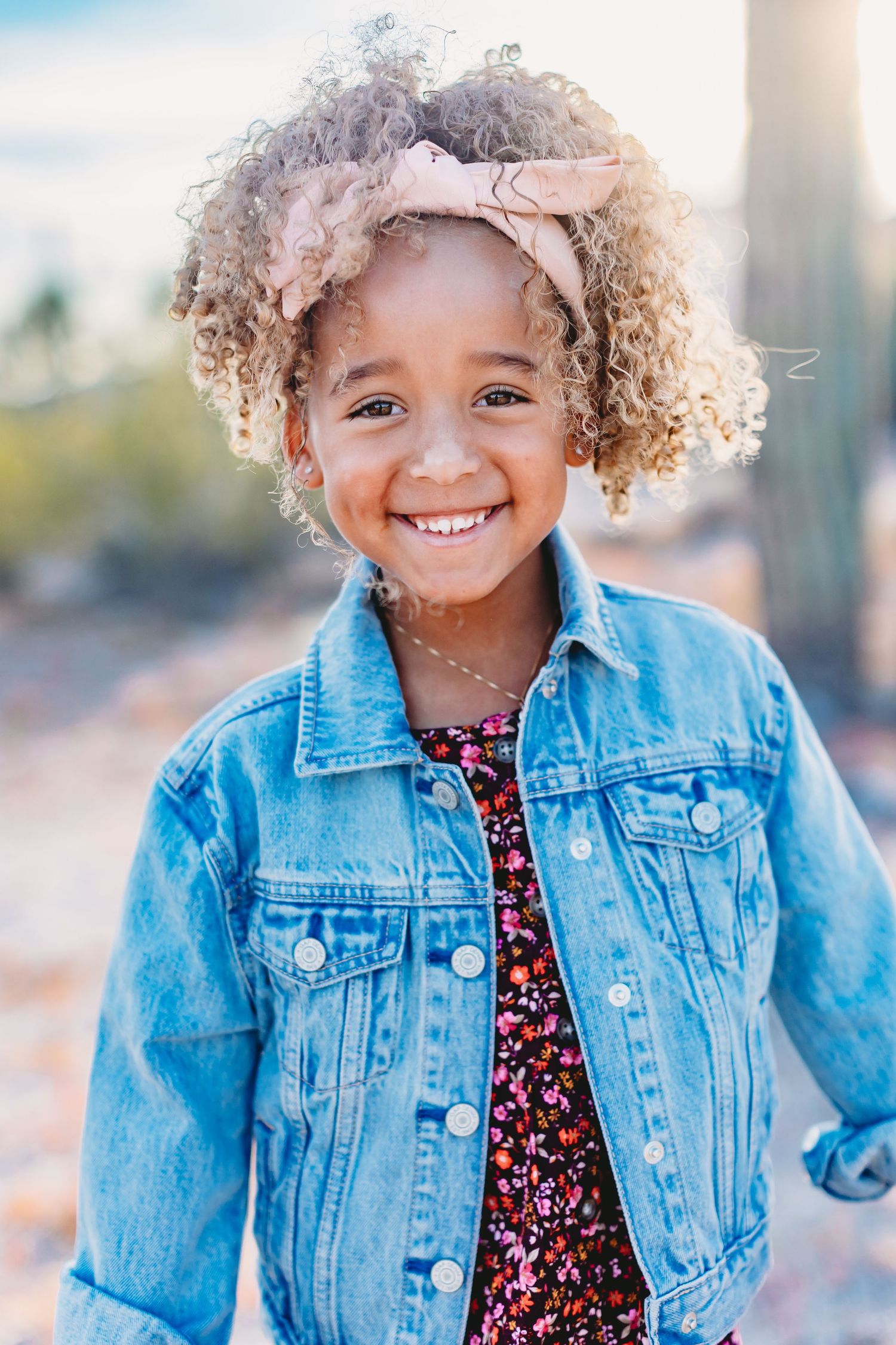 Child wearing denim jacket and floral dress poses with curly hair in desert setting.