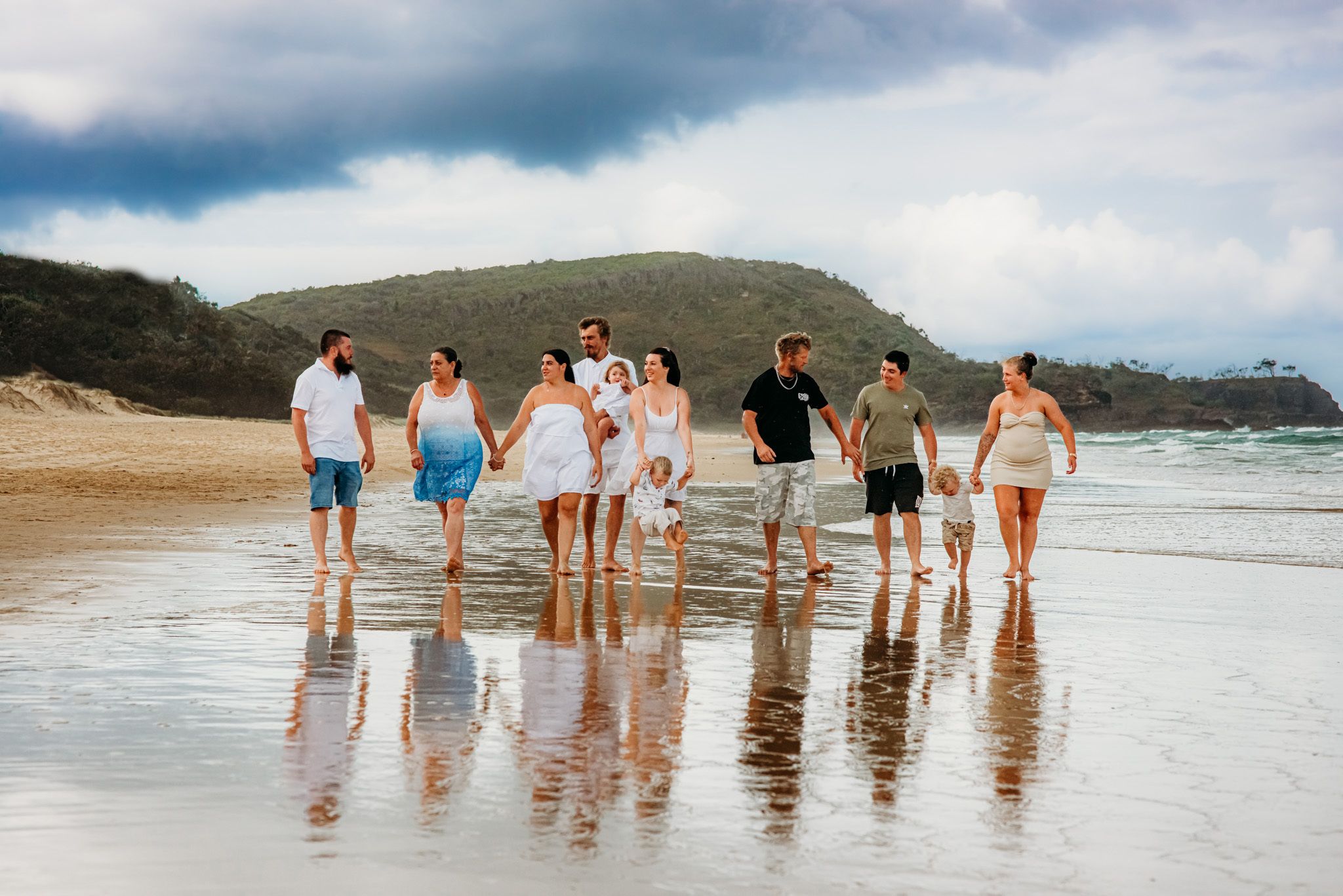 Line of people walking along beach shoreline with waves and cloudy sky.