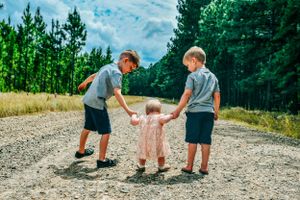 Family members interacting on a gravel path surrounded by green pine trees.