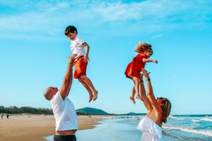 Two children being lifted playfully in the air at a sunny beach with ocean waves in the background.