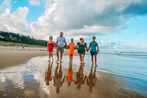 Group walking along a beach shoreline with reflections in wet sand under cloudy skies.