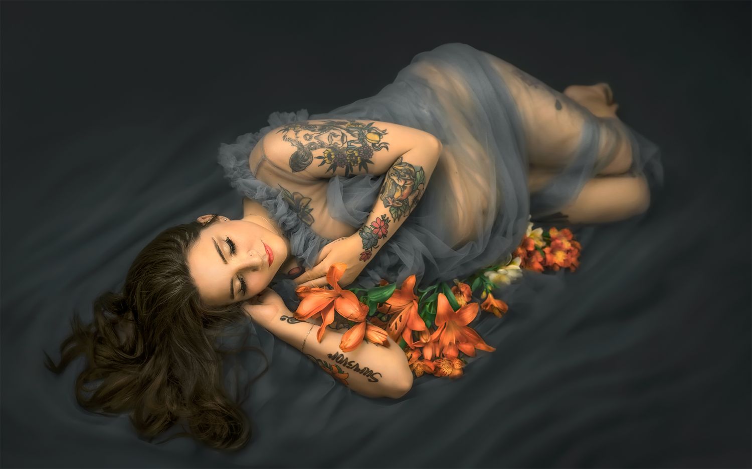 Artistic boudoir maternity portrait of a woman in sheer fabric lying with orange flowers in dark moody lighting.
