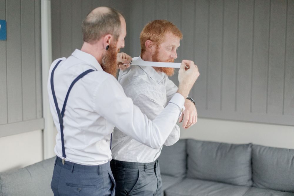 best man with ginger beard and suspenders helping bearded redhead groom get dressed