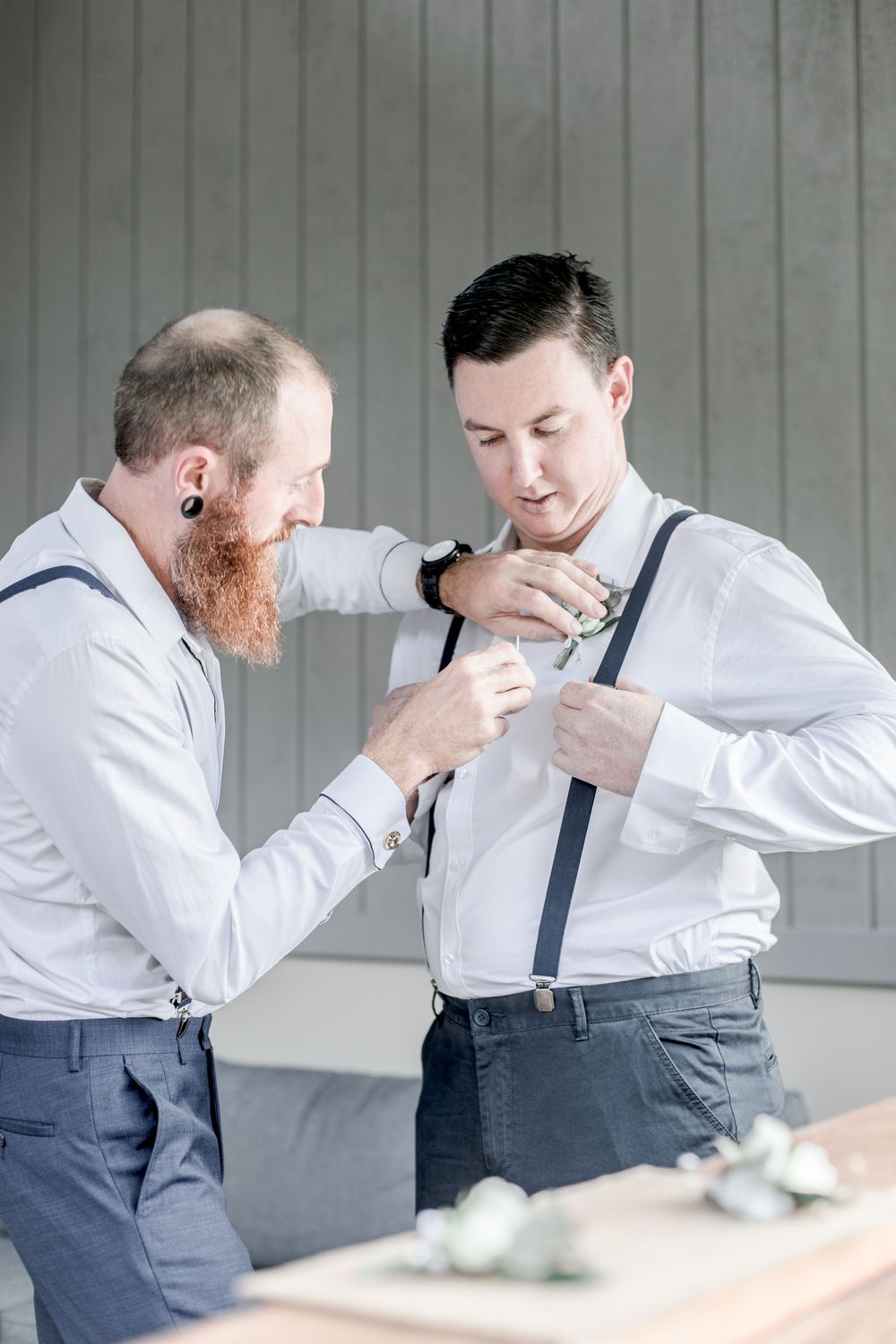 bearded best man in suspenders with ear stretchers helping clean shaven groomsman pin buttonhole to shirt