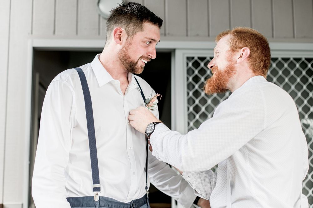 redhead groom wearing white shirt pinning white flower buttonhole onto bearded brunette groomsman wearing dark suspender