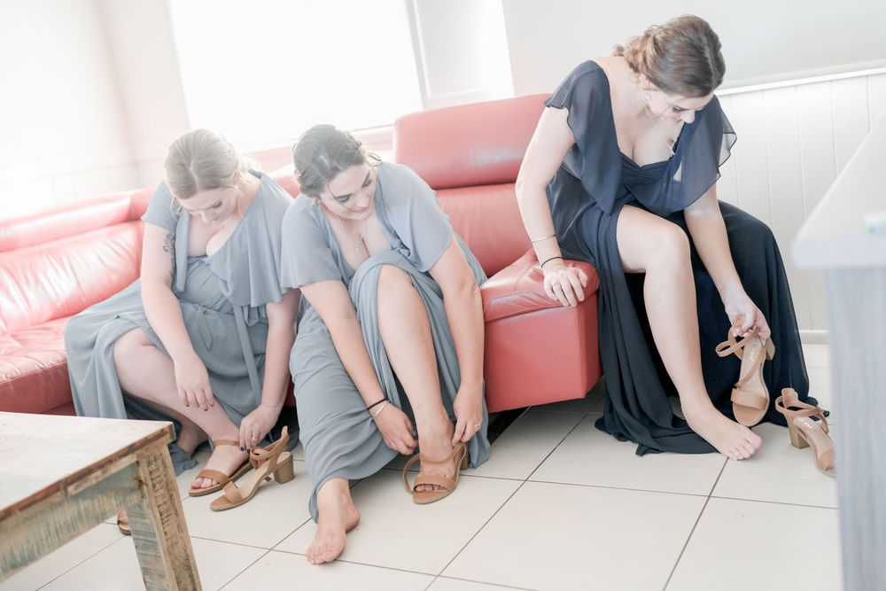 woman in dark blue long gown putting on shoes next to two women in sea blue long gowns sitting on red lounge