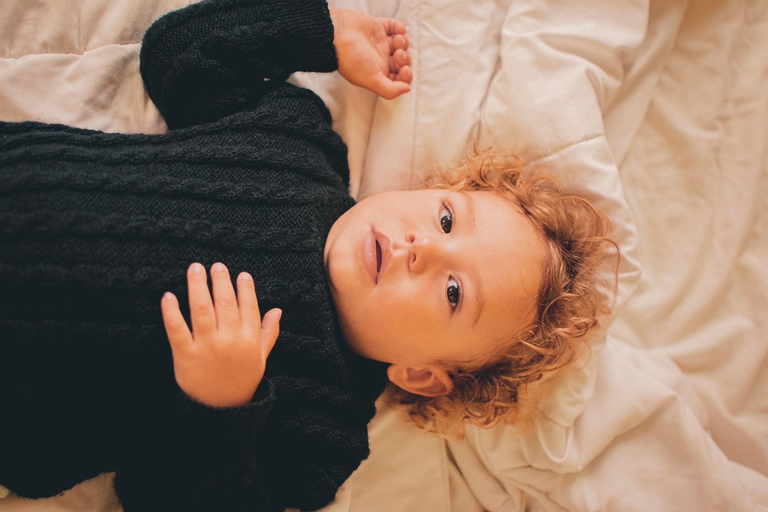Child with curly hair lying on cream colored bedding.
