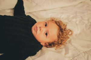 Close-up of child with curly blonde hair wearing black sweater lying on white bedding.