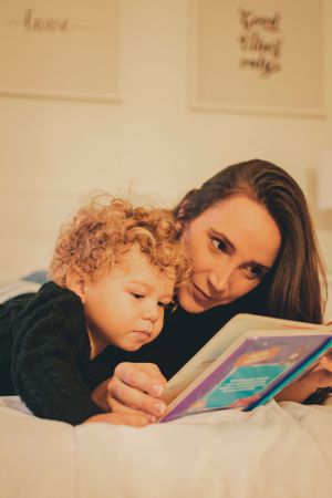 Parent and child reading a colorful book together while relaxing on white bedding.