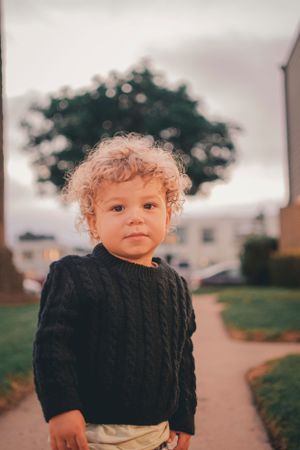 A young child with curly blonde hair wearing a black sweater stands on a path at dusk.