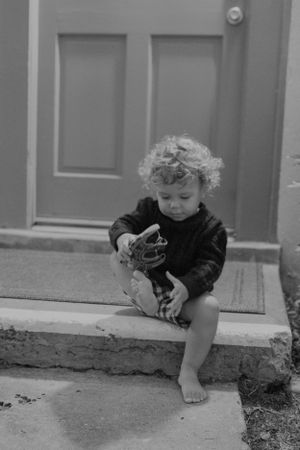 A small child in black clothing sits barefoot on a doorstep examining something in black and white.