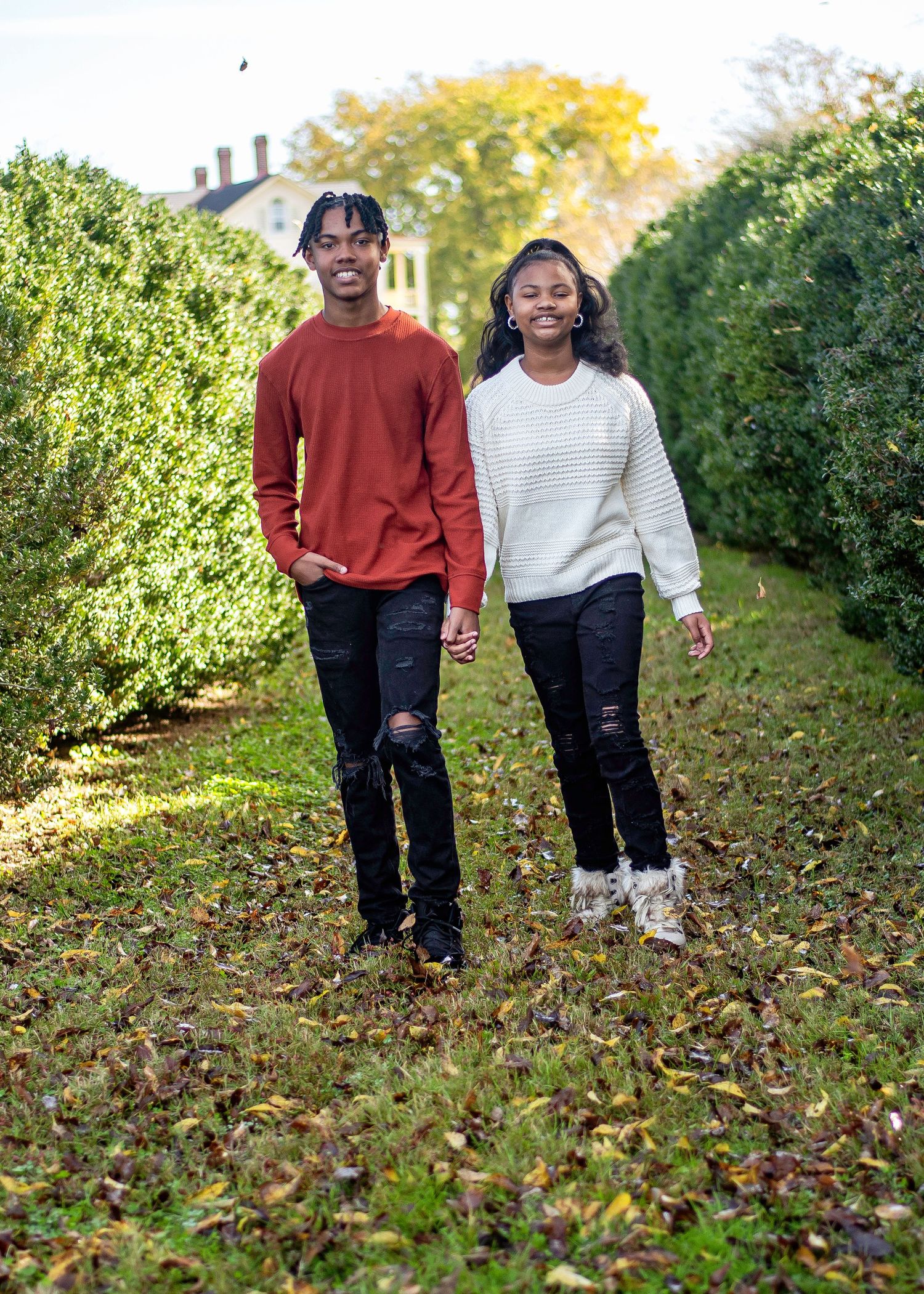 Two people walking together on a leaf-covered path between green hedges.