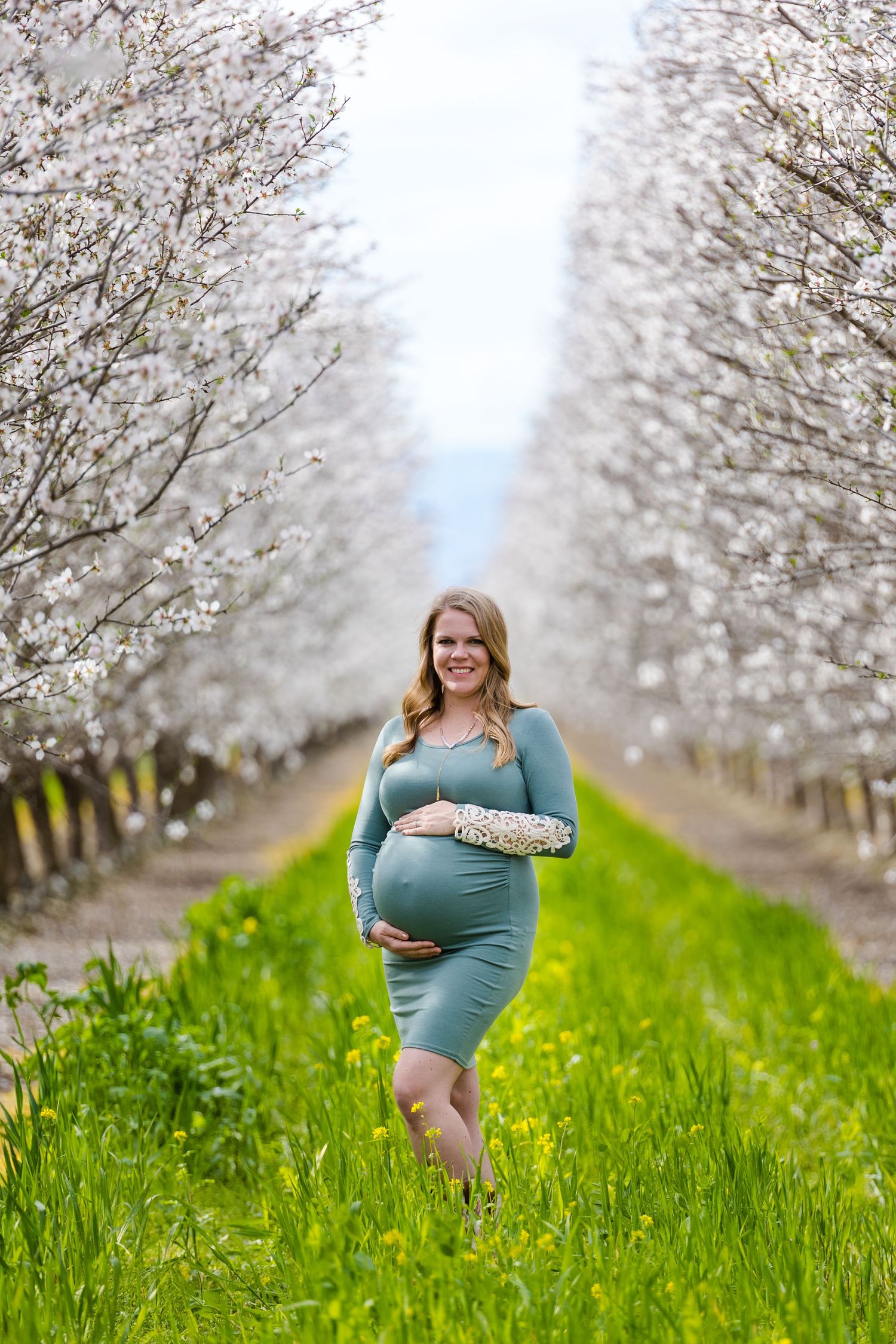 Woman in green grass in blooming almond orchard with mountain behind her in Maternity Session in Orland, California