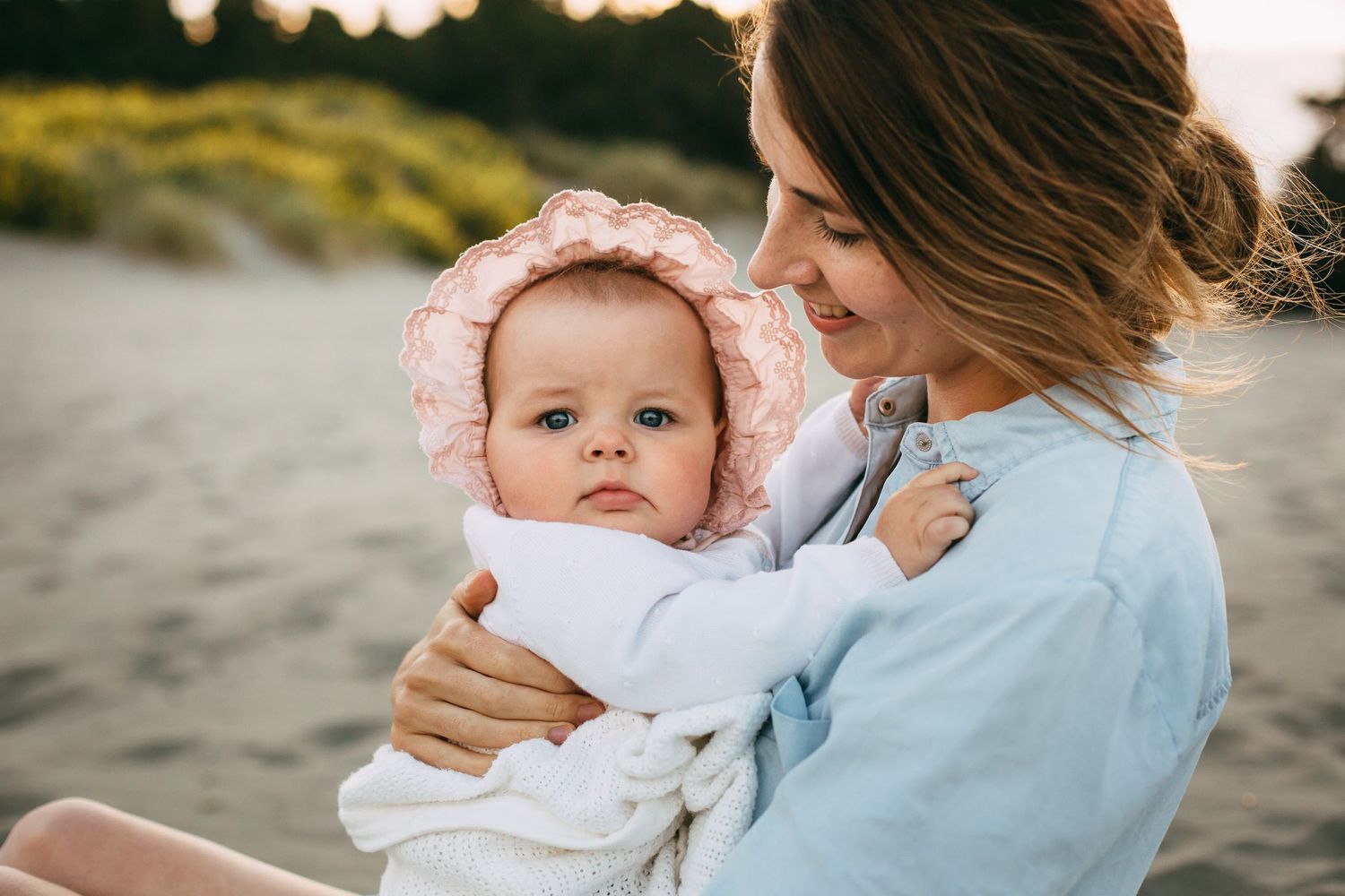 Mother holding her baby in a pink bonnet on the beach.