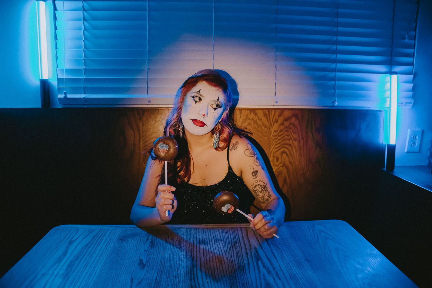 Dramatic portrait in blue lighting shows performer with white face paint sitting at a table.