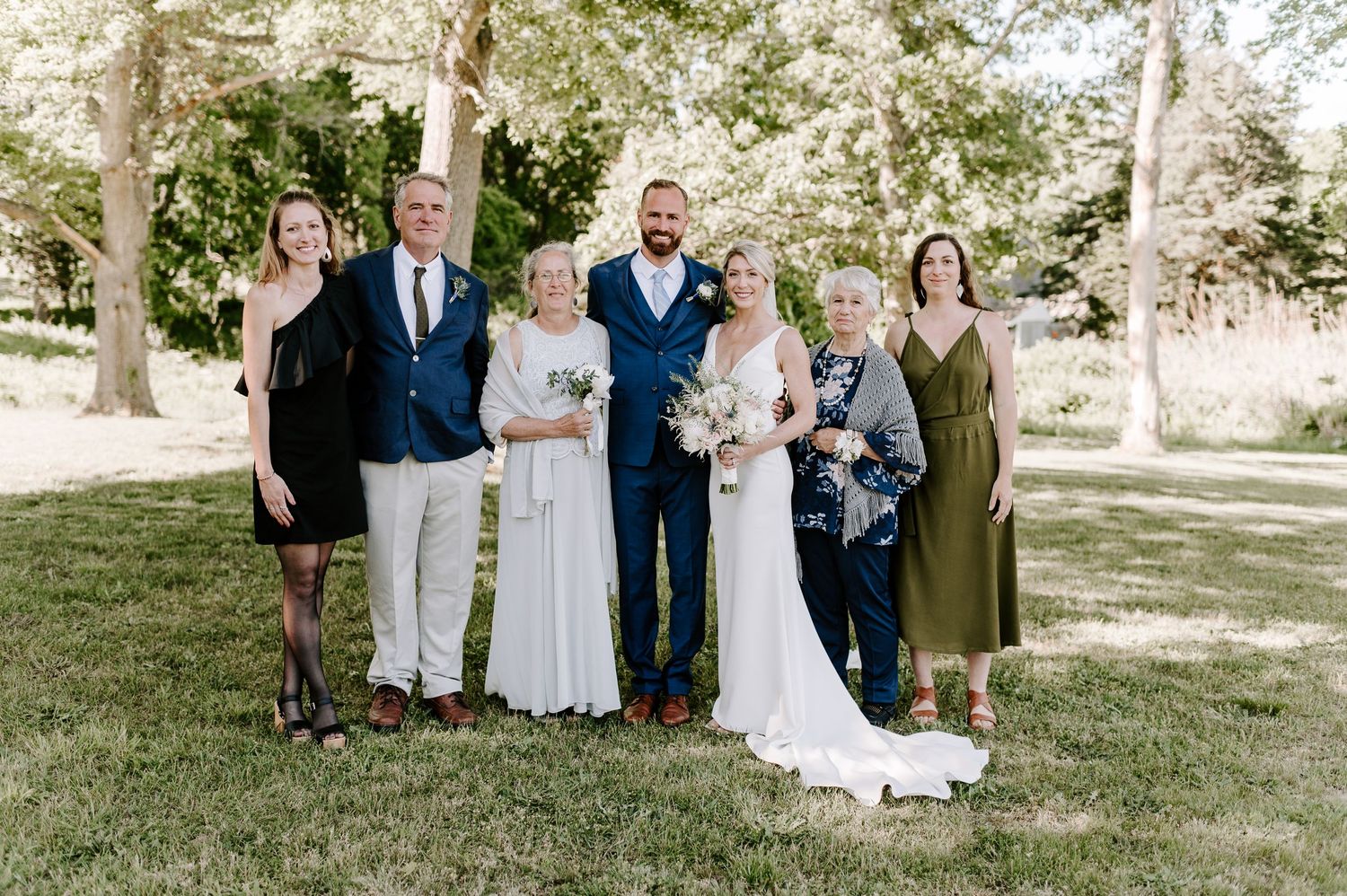 Wedding group photo taken outdoors under large trees in natural lighting.