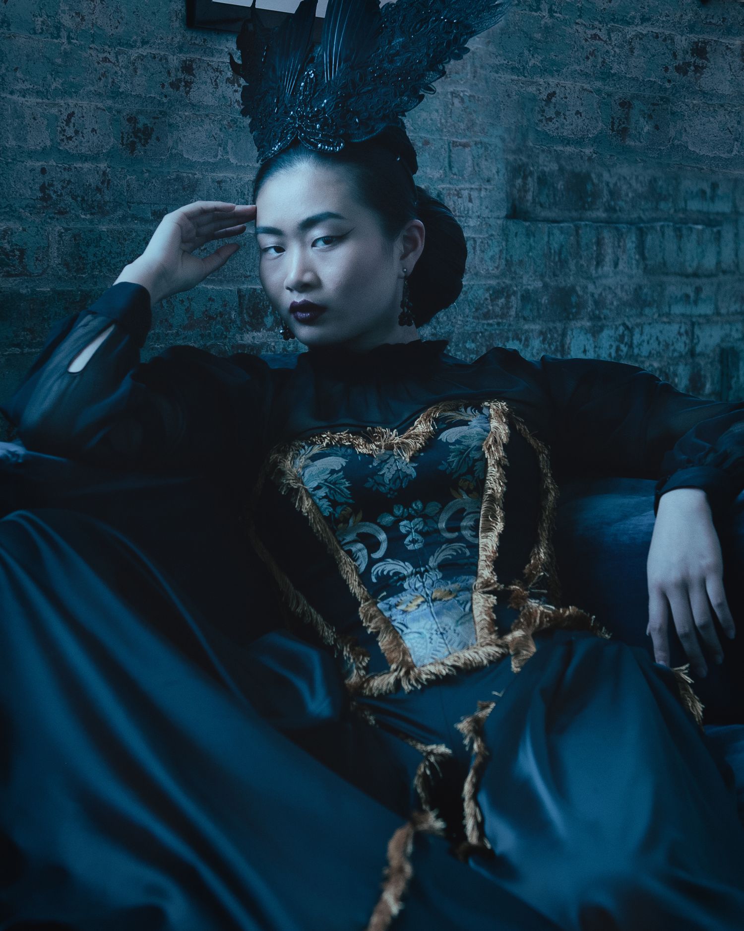 Model in dramatic dark baroque costume with ornate headpiece in moody setting.