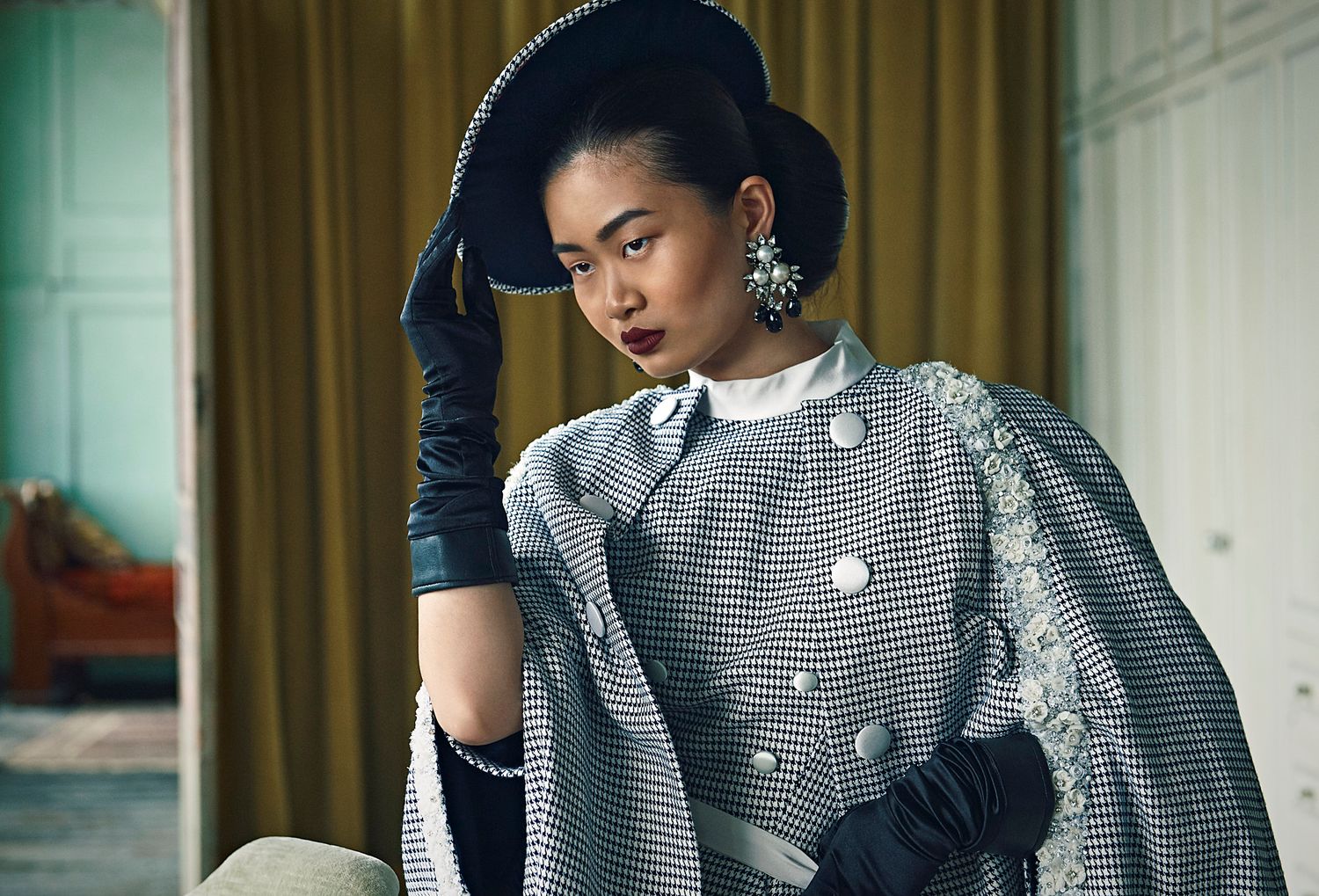 Elegant woman in vintage houndstooth cape and black gloves adjusting stylish hat.