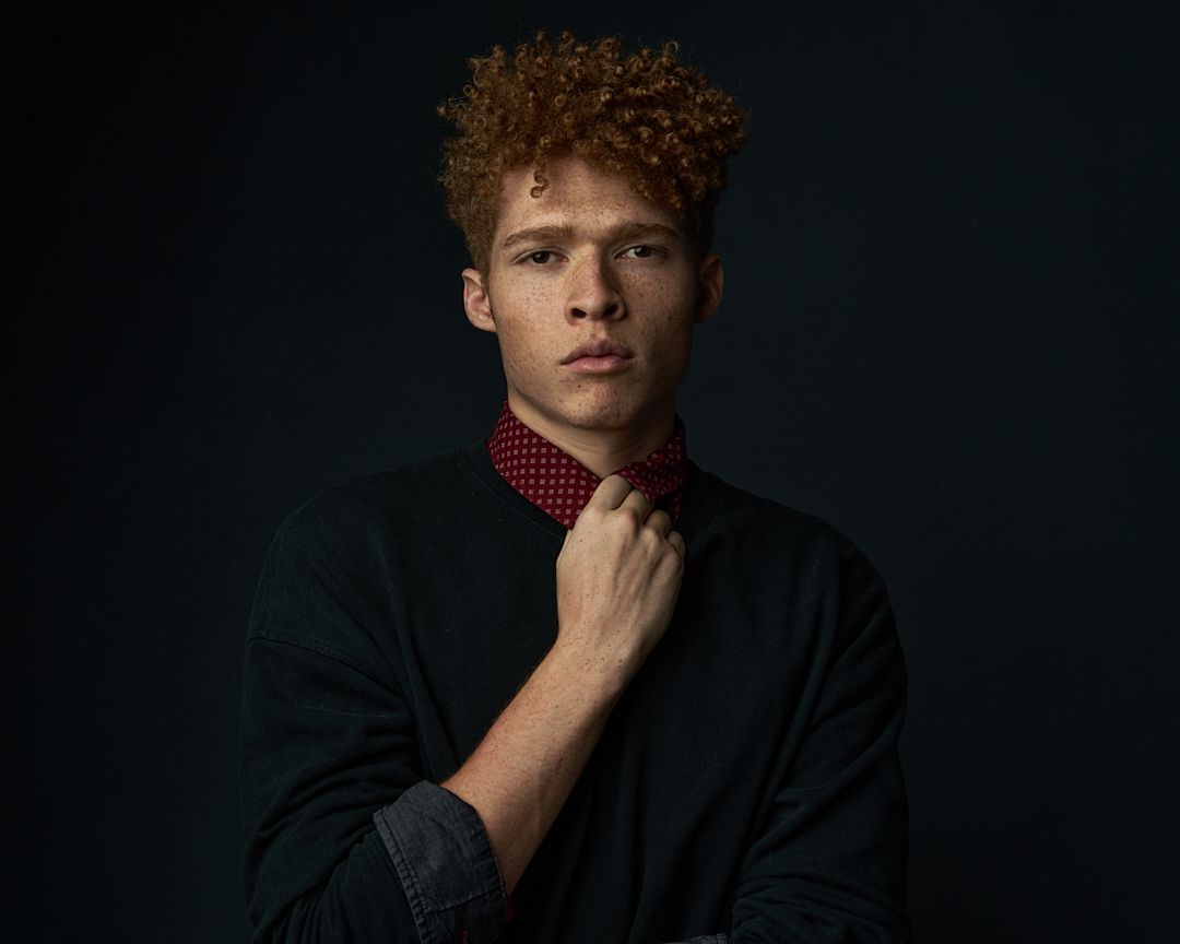 Young man with red curly hair adjusting red polka dot tie against dark background.