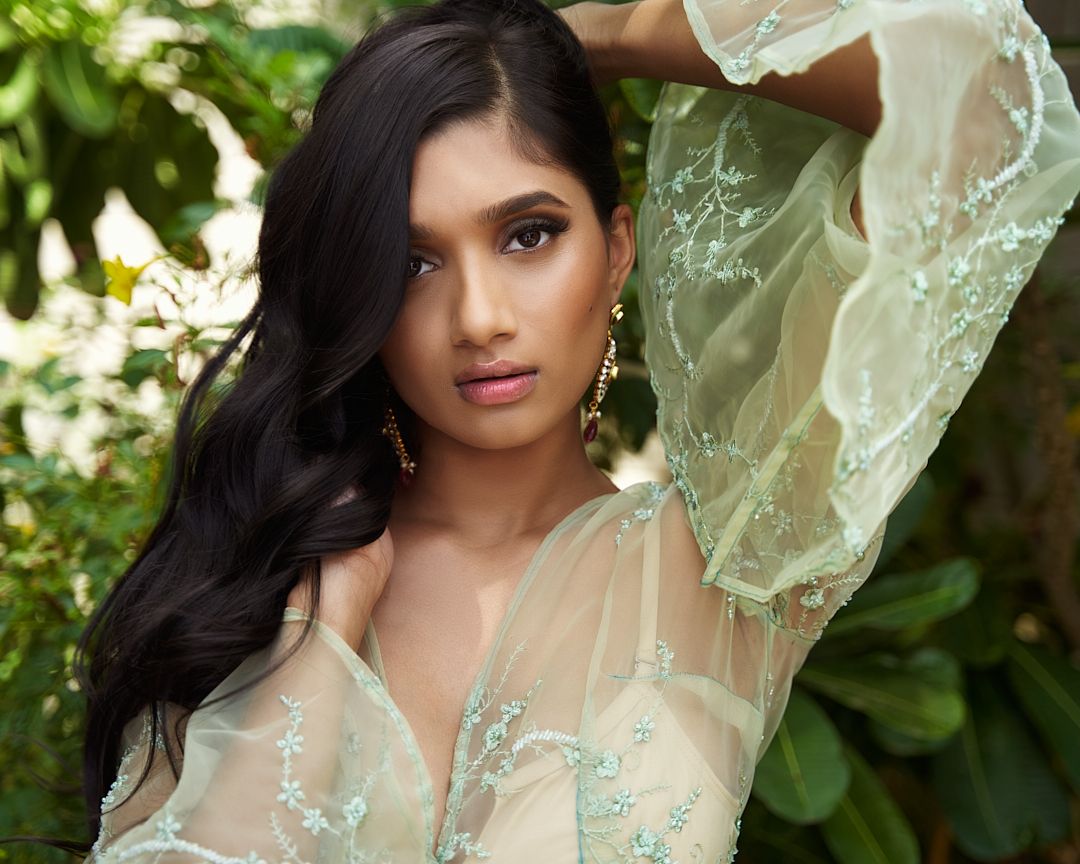 Young woman in sheer mint embroidered top posing outdoors near greenery.