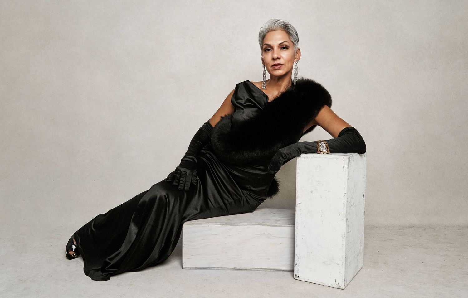 Elegant woman in black satin gown with fur trim and gloves posing on white block.