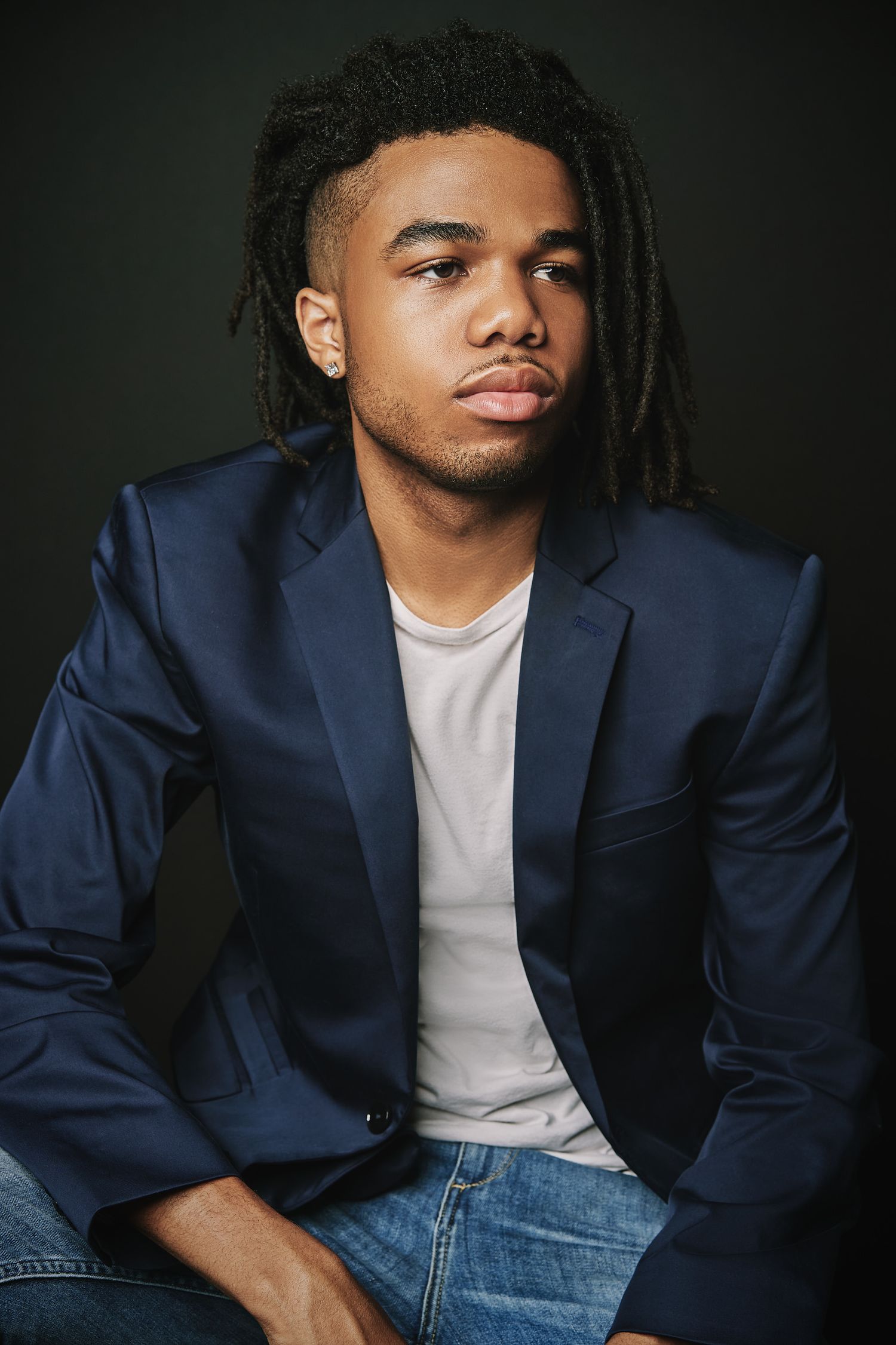 Stylish young man with dreadlocks wearing navy blazer and jeans against dark background.