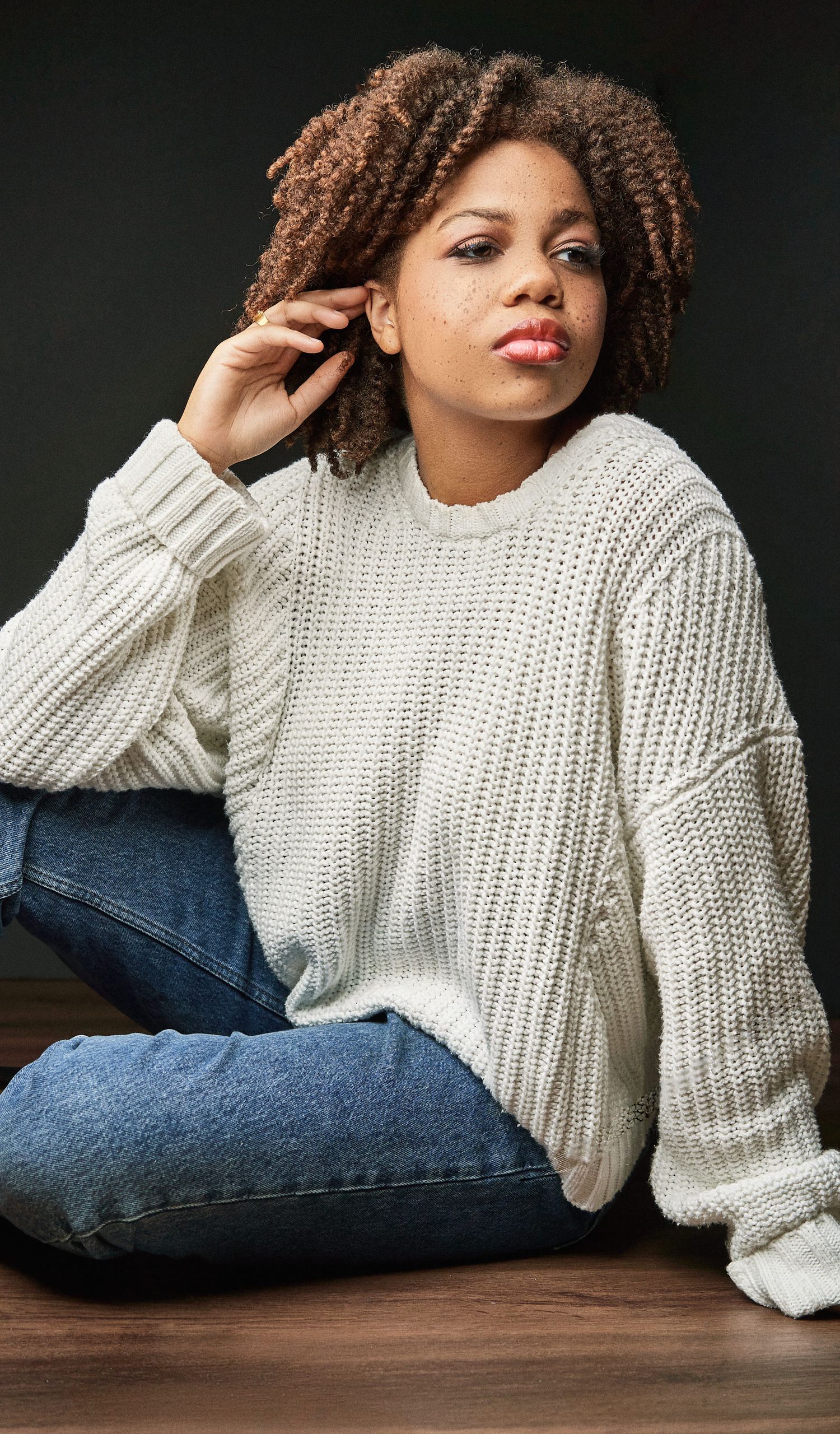 Young woman with curly hair in white knit sweater and jeans sitting on wooden floor.